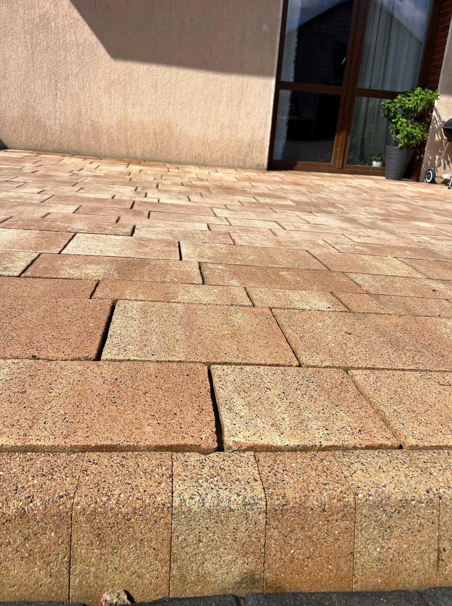 Czysta, brązowa kostka brukowa przed budynkiem z oknem, z doniczką. Widok z niskiej perspektywy podkreśla regularny wzór kostki i jej kolor.
