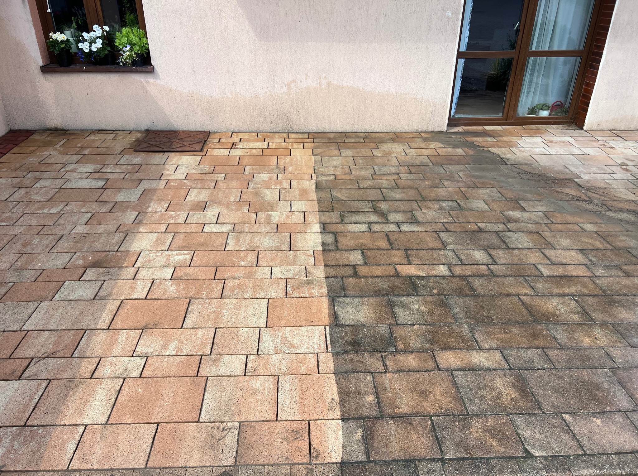 Kostka brukowa przed i po czyszczeniu, widoczna różnica w kolorze i czystości. Część powierzchni wyczyszczona, druga zabrudzona. Budynek w tle.