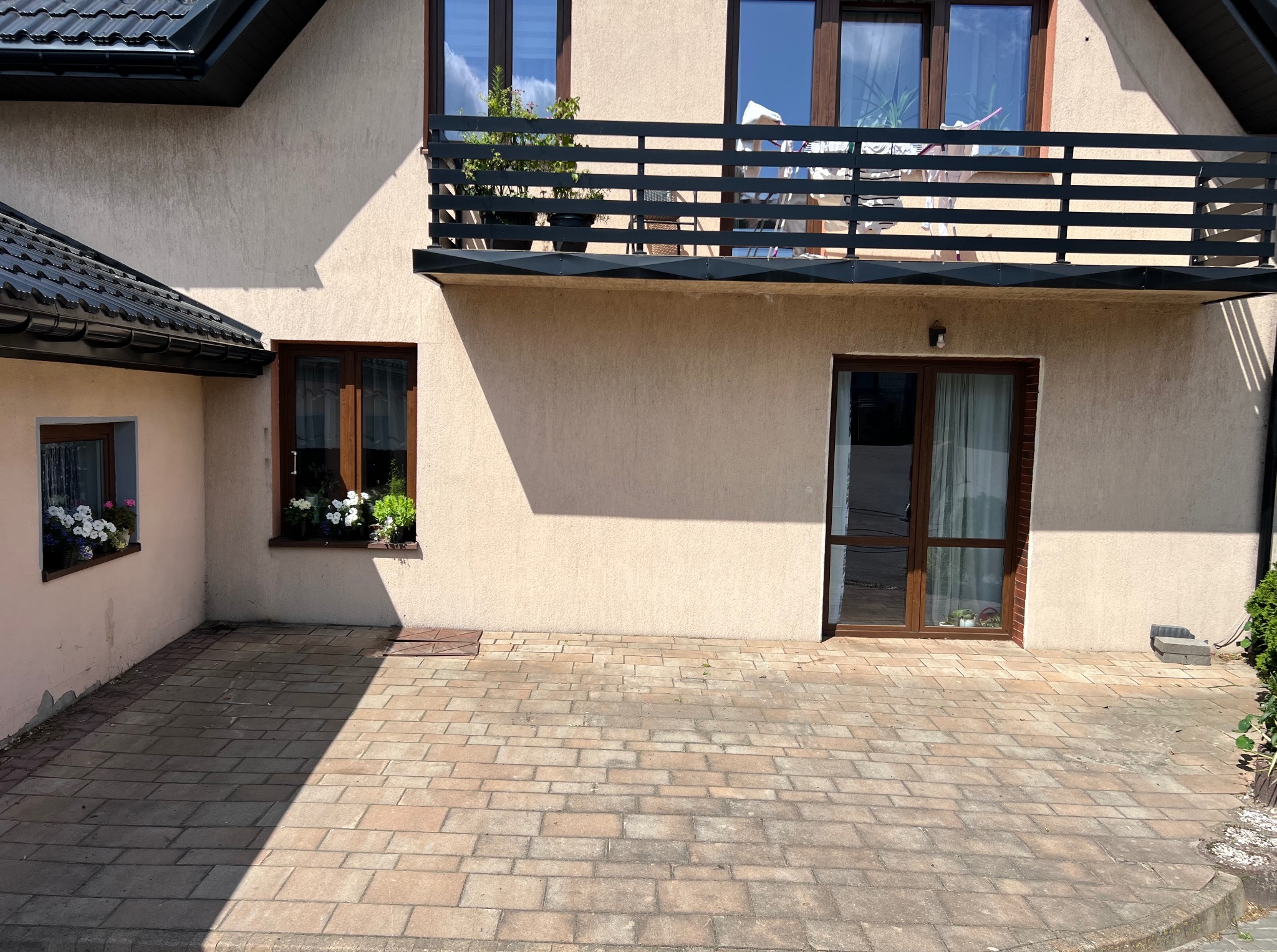 Dwukondygnacyjny dom z beżową elewacją, balkonem, brązowymi oknami i kostką brukową. Widoczne kwiaty w oknach i cień na kostce brukowej.