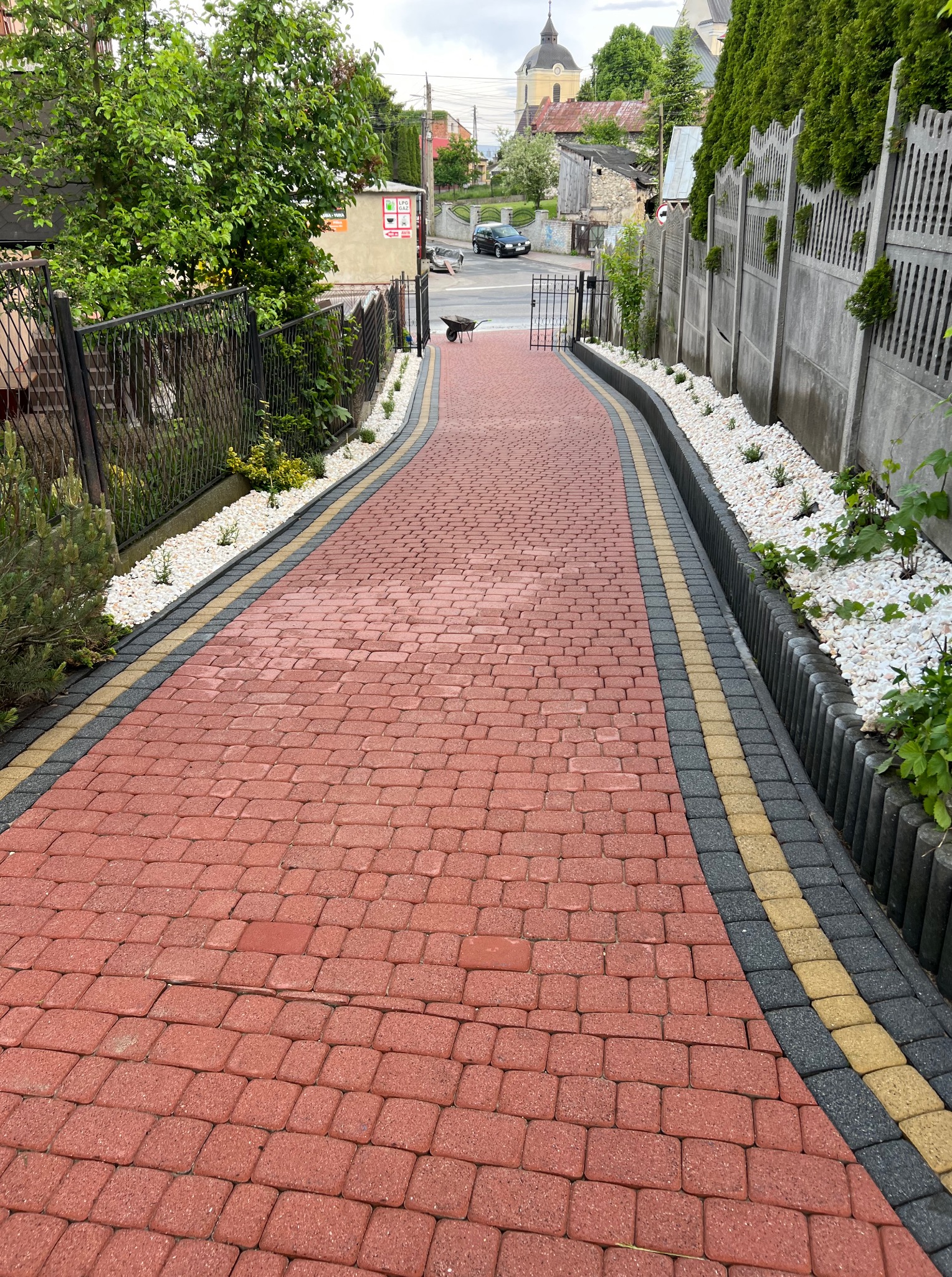 Czerwona kostka brukowa z obrzeżami w odcieniach szarości i żółci, otoczona białymi kamieniami i zielenią, prowadząca w dół w kierunku bramy i ulicy.