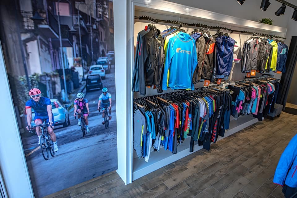 Wnętrze sklepu odzieżowego z odzieżą sportową na wieszakach i fototapetą przedstawiającą kolarzy. Widoczne kurtki, bluzy i koszulki rowerowe w różnych kolorach.