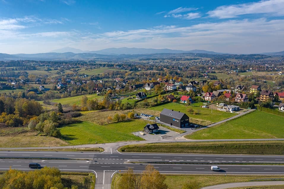 Panoramiczny widok z lotu ptaka na krajobraz z drogą, zabudowaniami i górami w tle pod błękitnym niebem.
