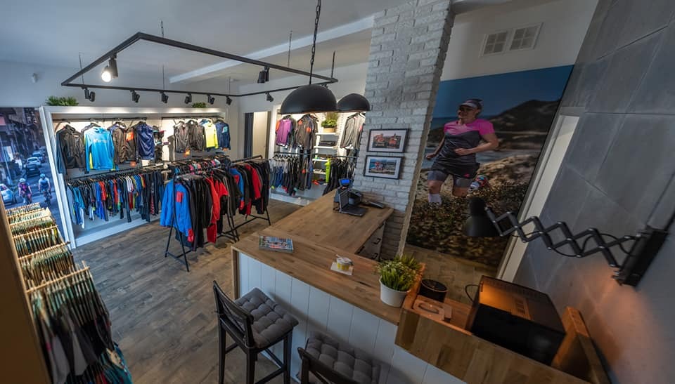 Wnętrze butiku odzieżowego z ekspozycją sportowej odzieży na wieszakach, drewnianą ladą i plakatem biegaczki na ścianie. Stylowe oświetlenie i nowoczesny design.