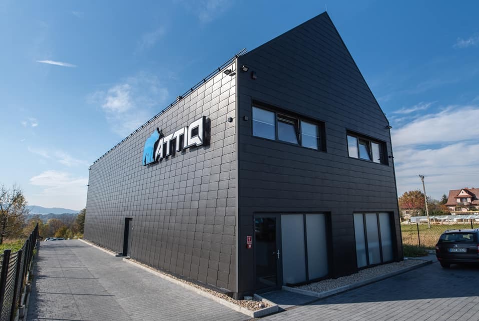 Nowoczesny budynek firmy odzieżowej z ciemną elewacją, logo i dużymi oknami, otoczony zielenią i brukowanym podjazdem w słoneczny dzień.