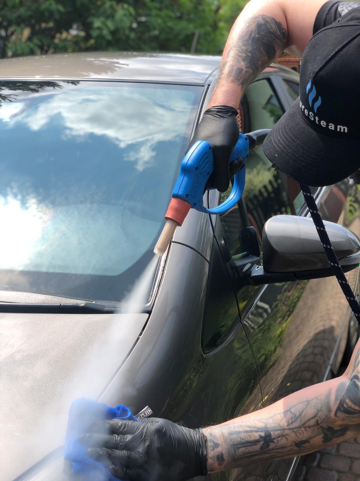 Czyszczenie parowe auta: mężczyzna w czarnych rękawicach i czapce z logo czyści szary samochód parownicą i niebieską ściereczką z mikrofibry.