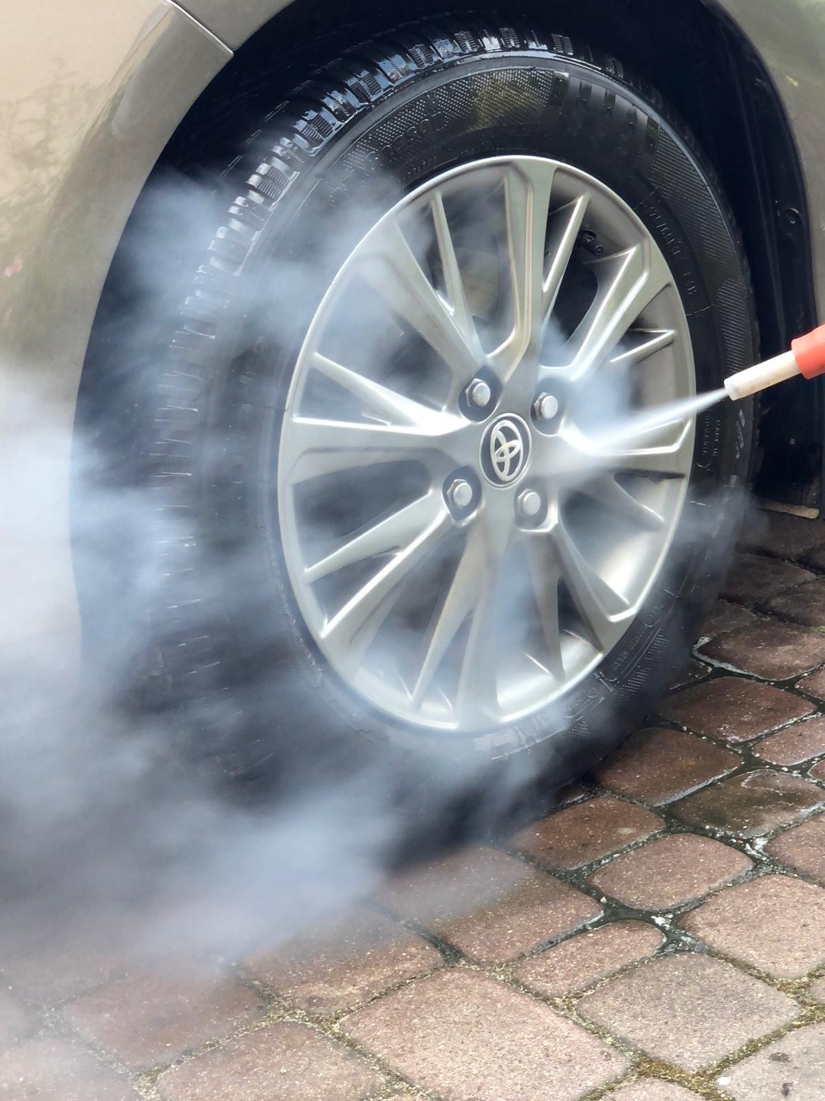 Czyszczenie felgi aluminiowej Toyoty myjką ciśnieniową parową na kostce brukowej. Widoczny strumień pary pod ciśnieniem i fragment opony.