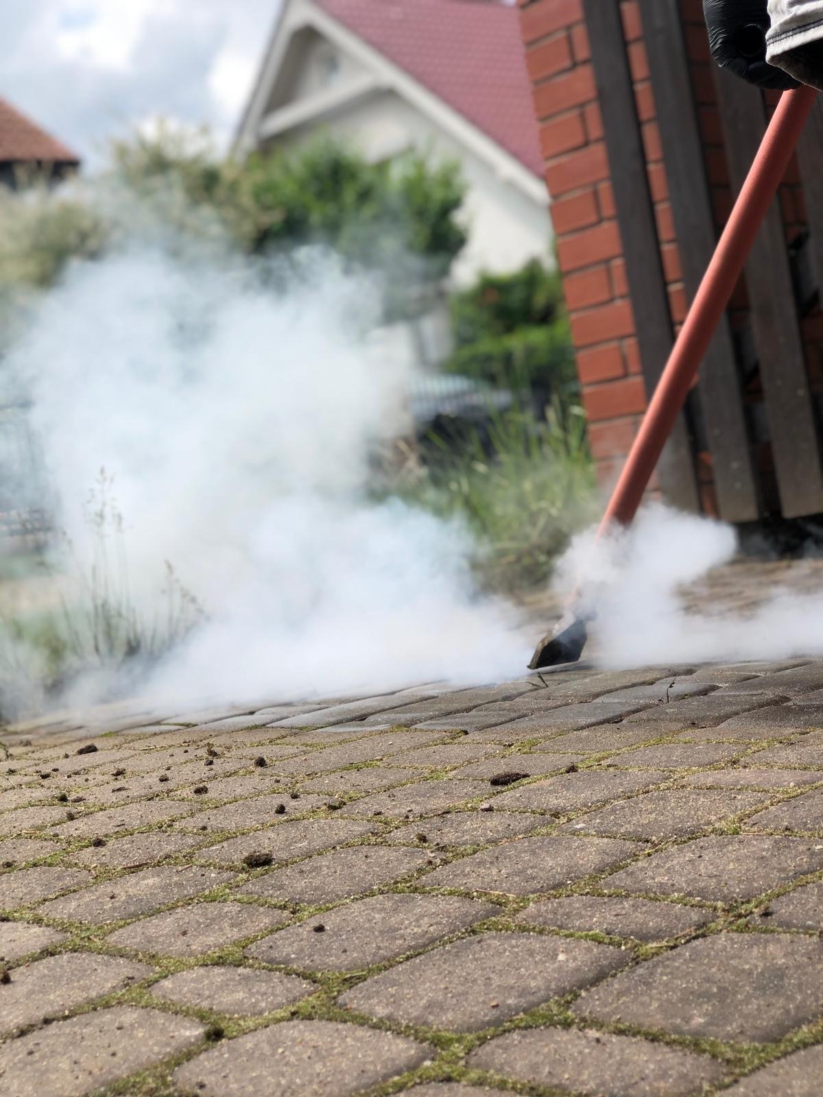 Czyszczenie kostki brukowej parą wodną. Z bliska widoczna dysza parowa i unosząca się para. W tle dom z czerwoną dachówką i zielona roślinność.