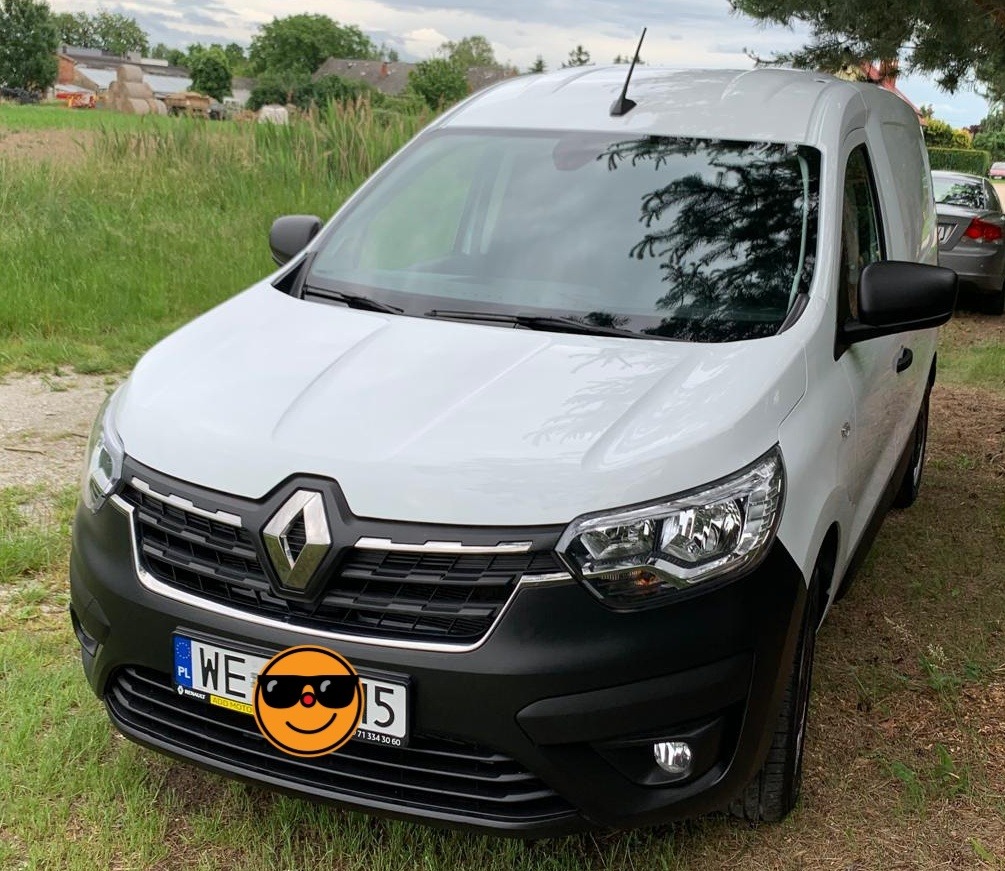 Biały Renault Kangoo Van z anteną, zaparkowany na trawie z zamazaną tablicą rejestracyjną, sugerujący mobilne usługi myjni lub transport tapicerki.