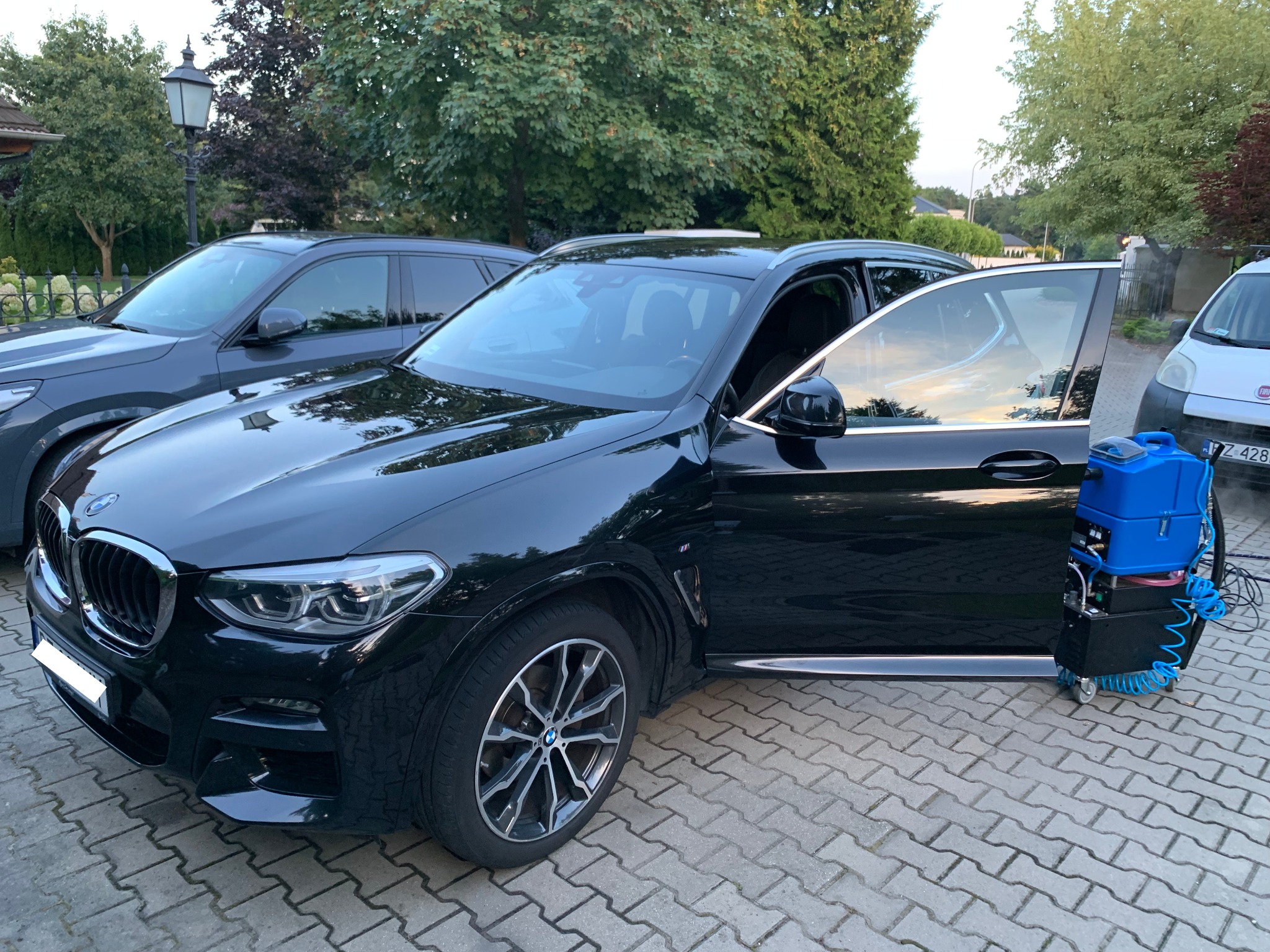 Czarny BMW X3 z otwartymi drzwiami pasażera i sprzętem do prania tapicerki obok. Usługa prania tapicerki samochodowej na zewnątrz.