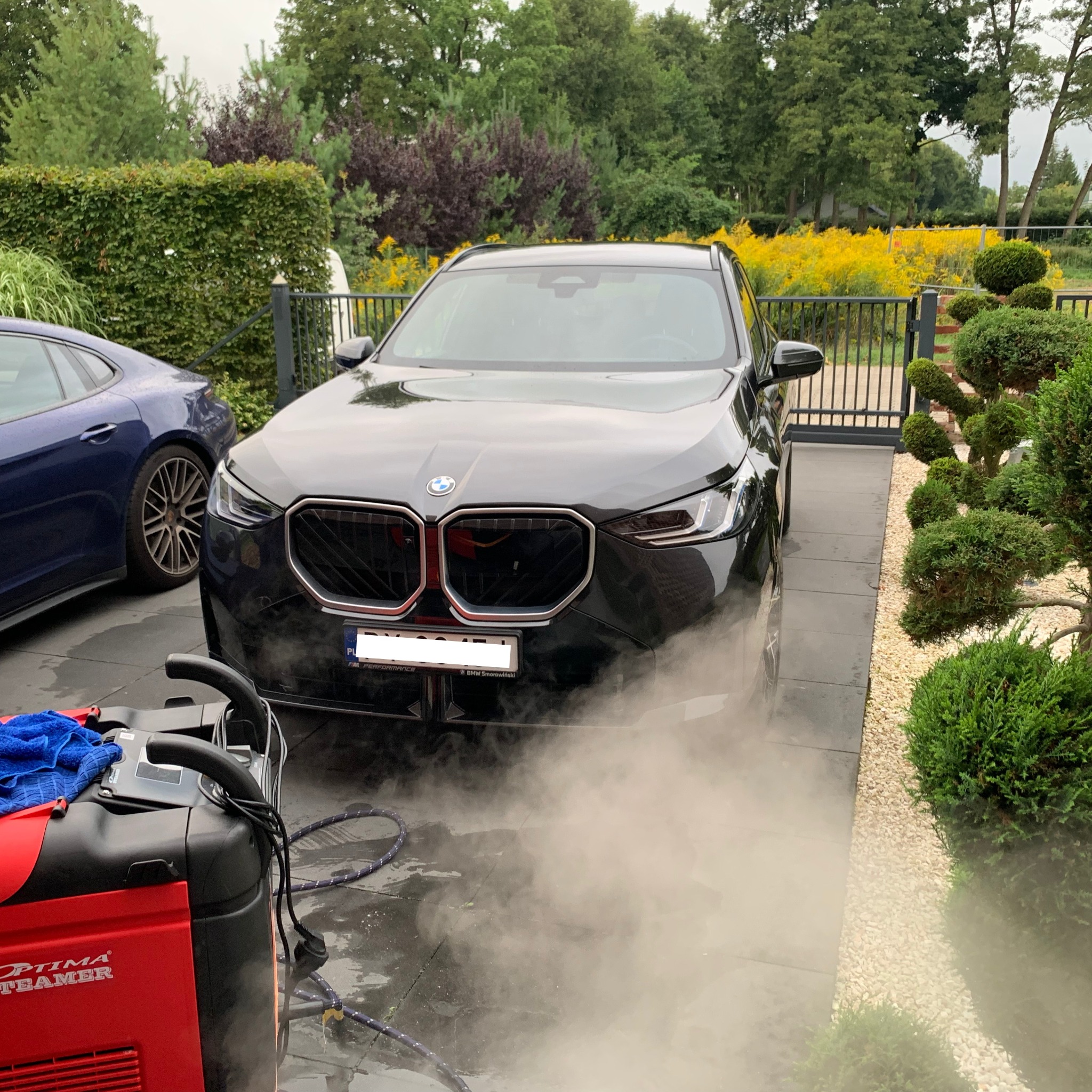 Czarny samochód BMW myty parą na posesji z zadbanym ogrodem. Widoczna maszyna parowa Optima Steamer i fragment drugiego auta.