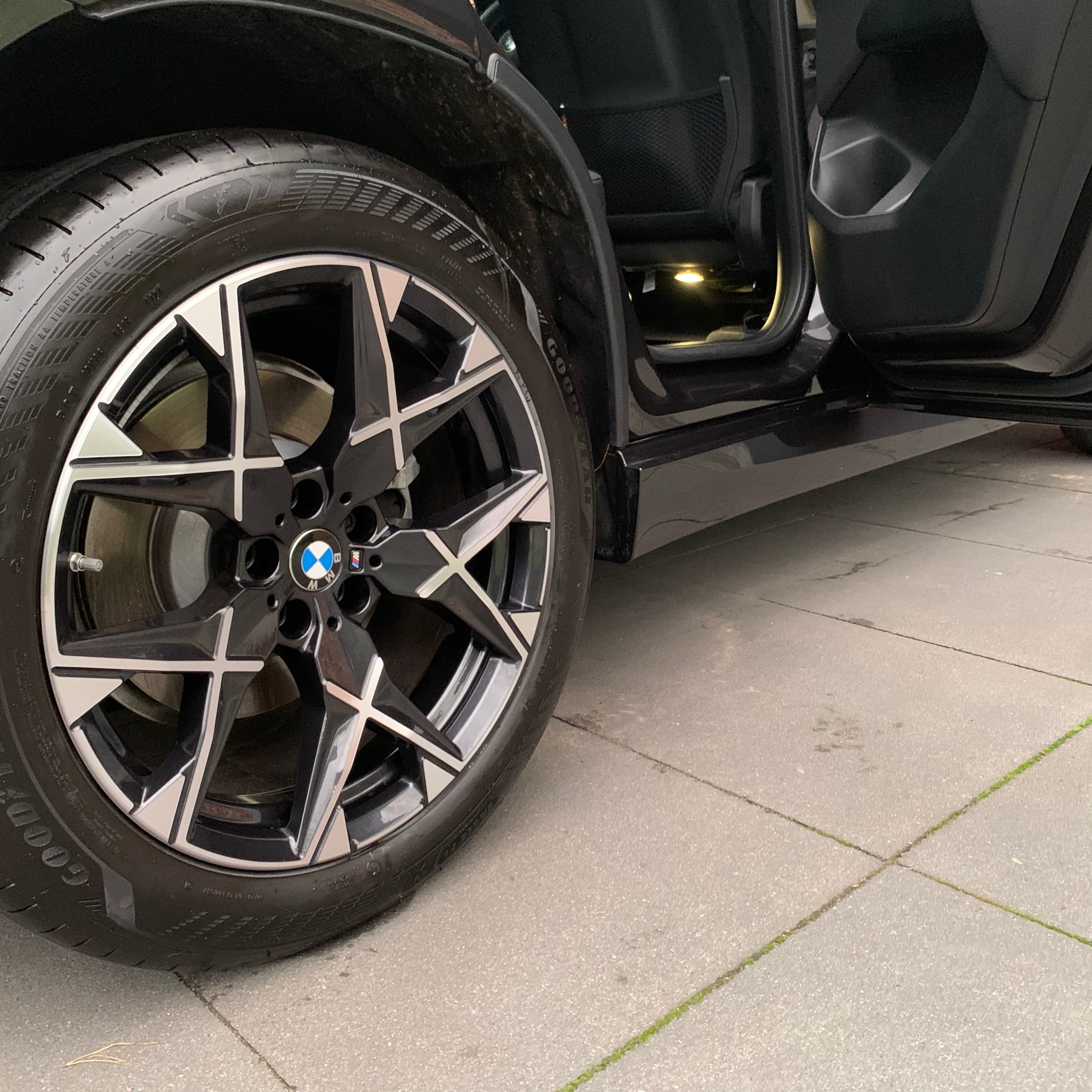 Błyszczące, czyste koło BMW z aluminiową felgą na tle otwartych drzwi samochodu. Widoczne oświetlenie wnętrza i fragment chodnika.