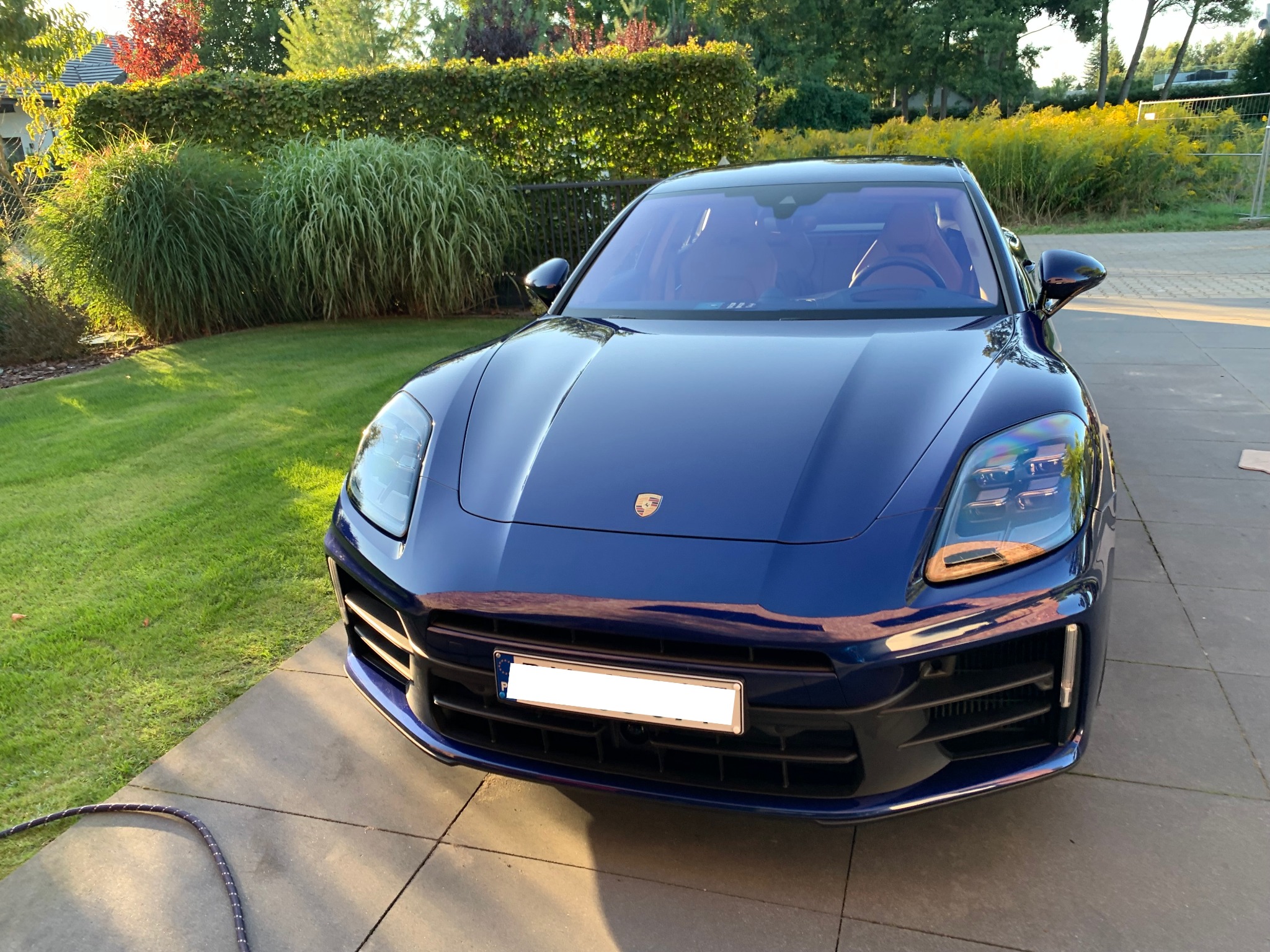 Błyszczący, granatowy samochód osobowy marki Porsche zaparkowany na kostce brukowej, z widocznym wężem i zielenią w tle.