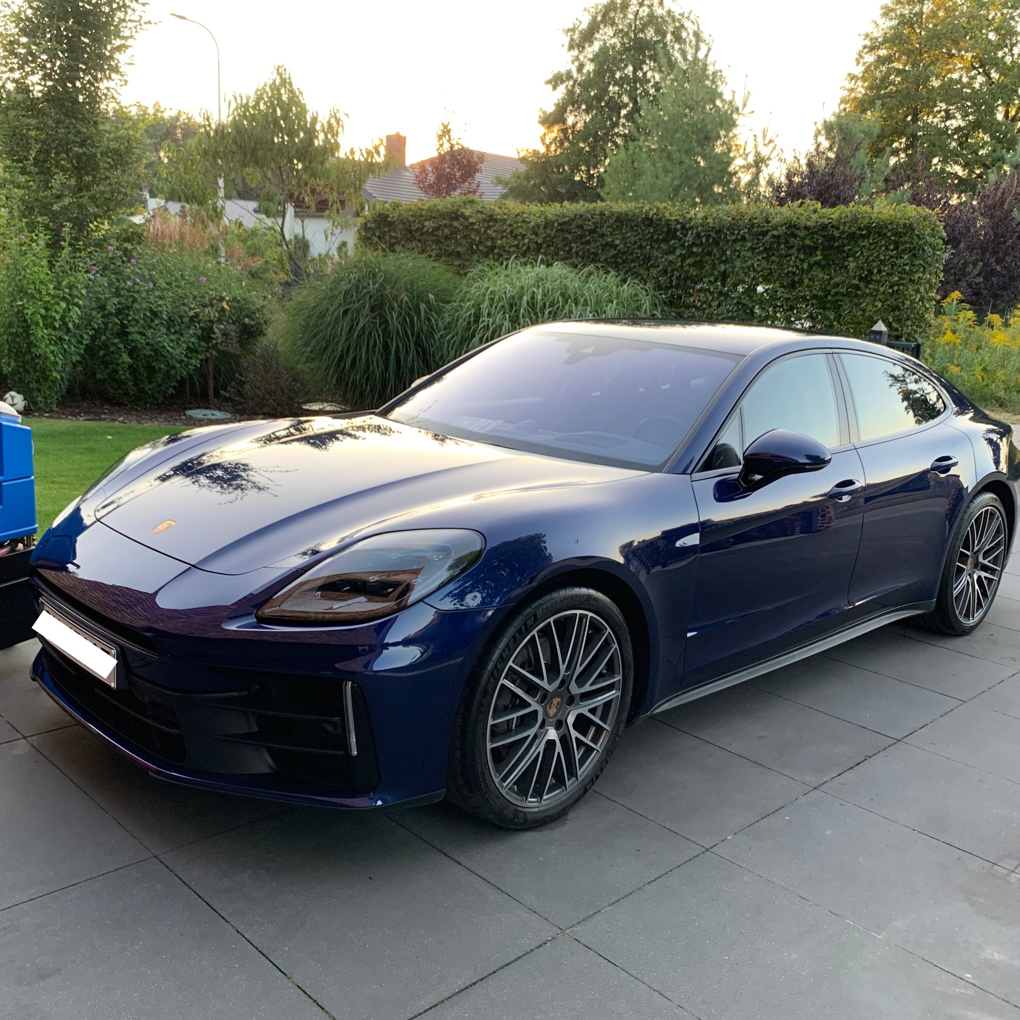 Błyszczący, granatowy Porsche Panamera po myciu, zaparkowany na szarym bruku z zielenią w tle. Widoczne detale felg i odbicia.