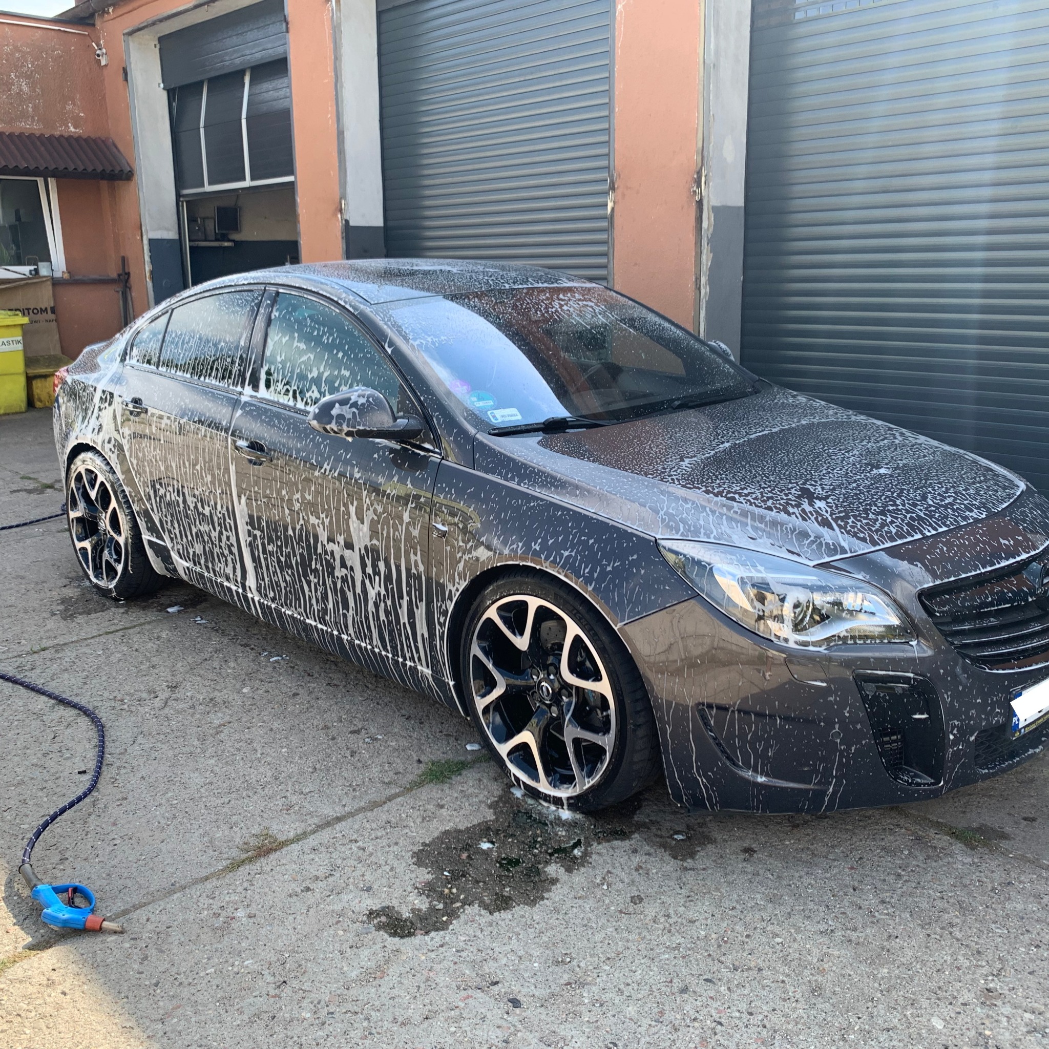 Szary samochód osobowy pokryty pianą podczas mycia, widok z boku na tle szarej bramy garażowej i fragmentu budynku. Wąż do mycia leży na ziemi.