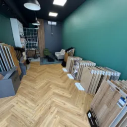 Salon z ekspozycją paneli podłogowych ułożonych w jodełkę, z widocznymi próbkami różnych producentów, oświetleniem sufitowym i elementami wystroju wnętrz.