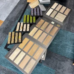 Katalog z próbkami paneli podłogowych i wykładzin na stoliku z metalową ramą, na tle dywanu w geometryczne wzory. Prezentacja różnych wzorów i kolorów.