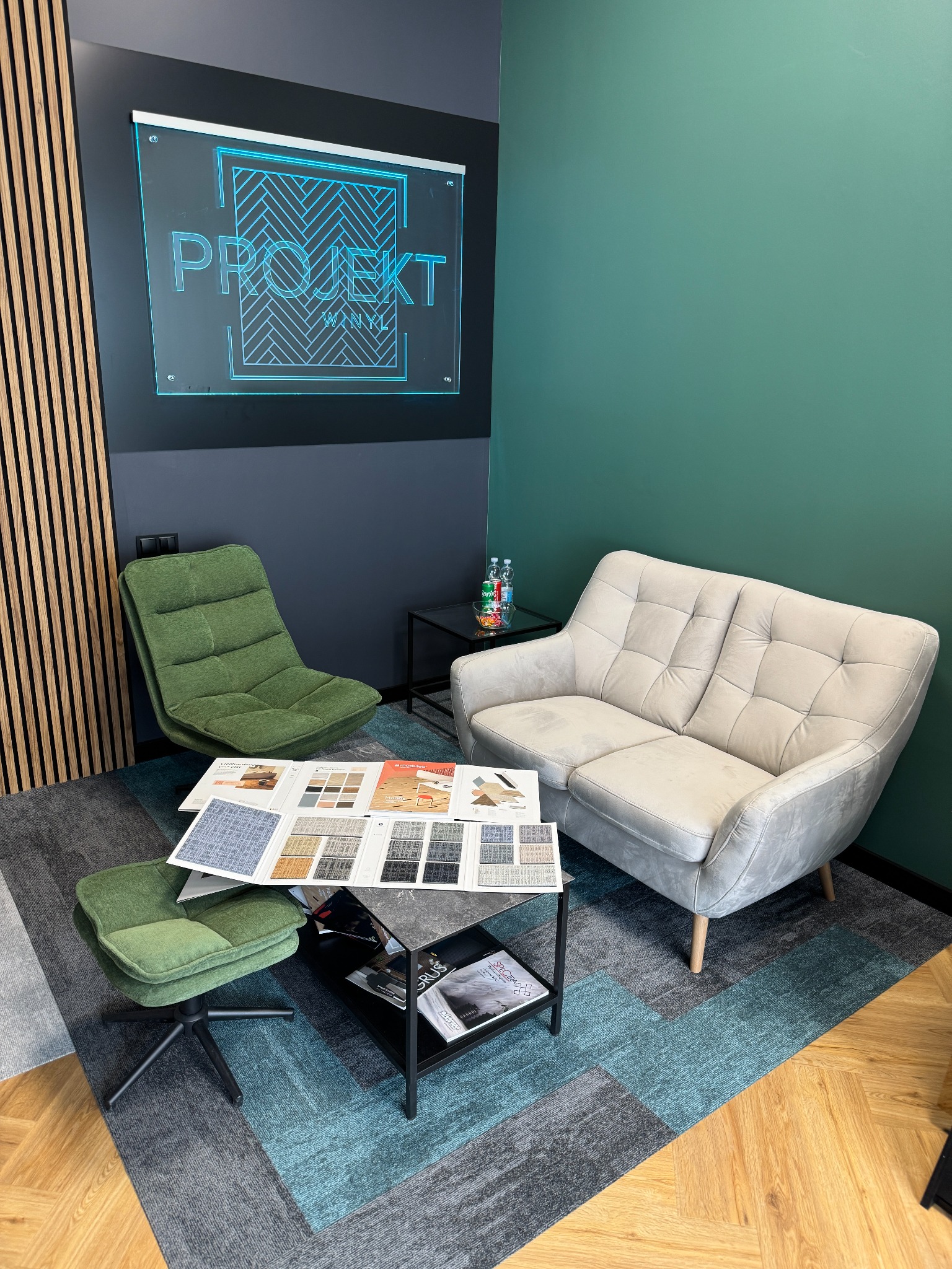 Salon z próbkami wykładzin dywanowych na stoliku, zielonym fotelem i szarą sofą. Na ścianie dekoracyjny panel drewniany i podświetlany napis Projekt Winyl.