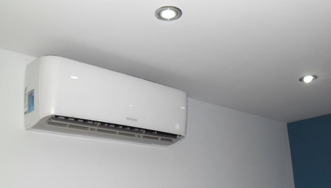 Biała klimatyzacja marki Samsung zamontowana na ścianie w pomieszczeniu z białym sufitem i dwoma punktowymi źródłami światła.