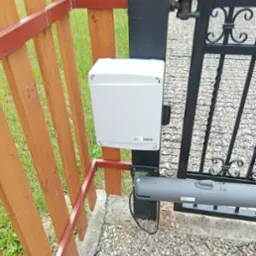 Zamontowany napęd bramy skrzydłowej z białą obudową sterującą na czarnym słupku. Widoczny fragment drewnianego ogrodzenia i podłoże z kamyków.