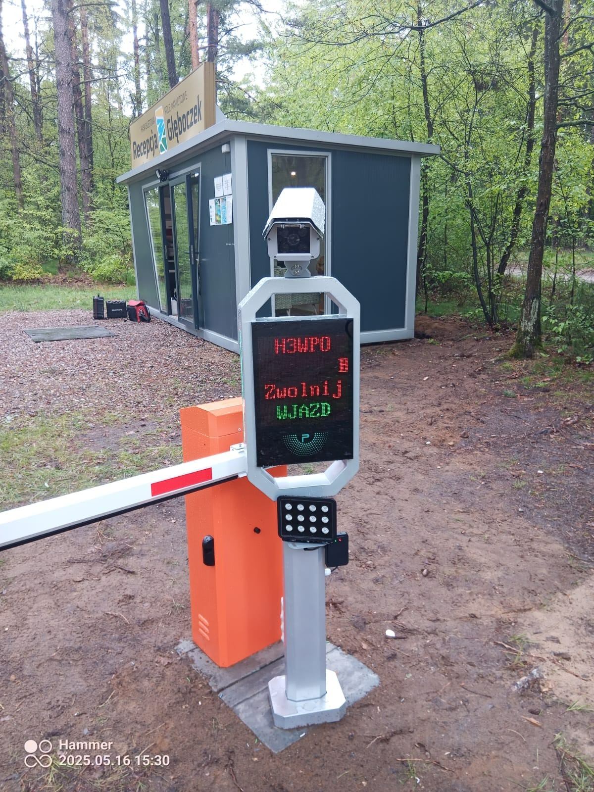 Szlaban automatyczny z kamerą i wyświetlaczem LED 'Zwolnij Wjazd' przy budce recepcji, teren leśny.
