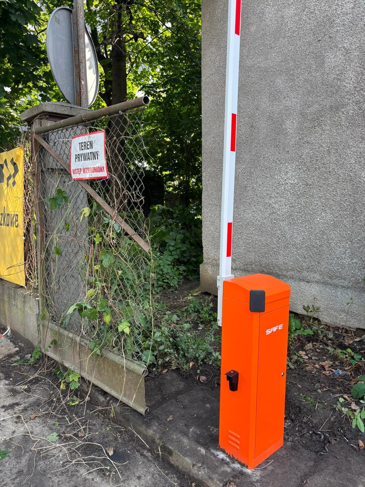 Pomarańczowa automatyczna bariera wjazdowa SAFE przy zarośniętym ogrodzeniu z tabliczką 'Teren prywatny. Wstęp wzbroniony'.
