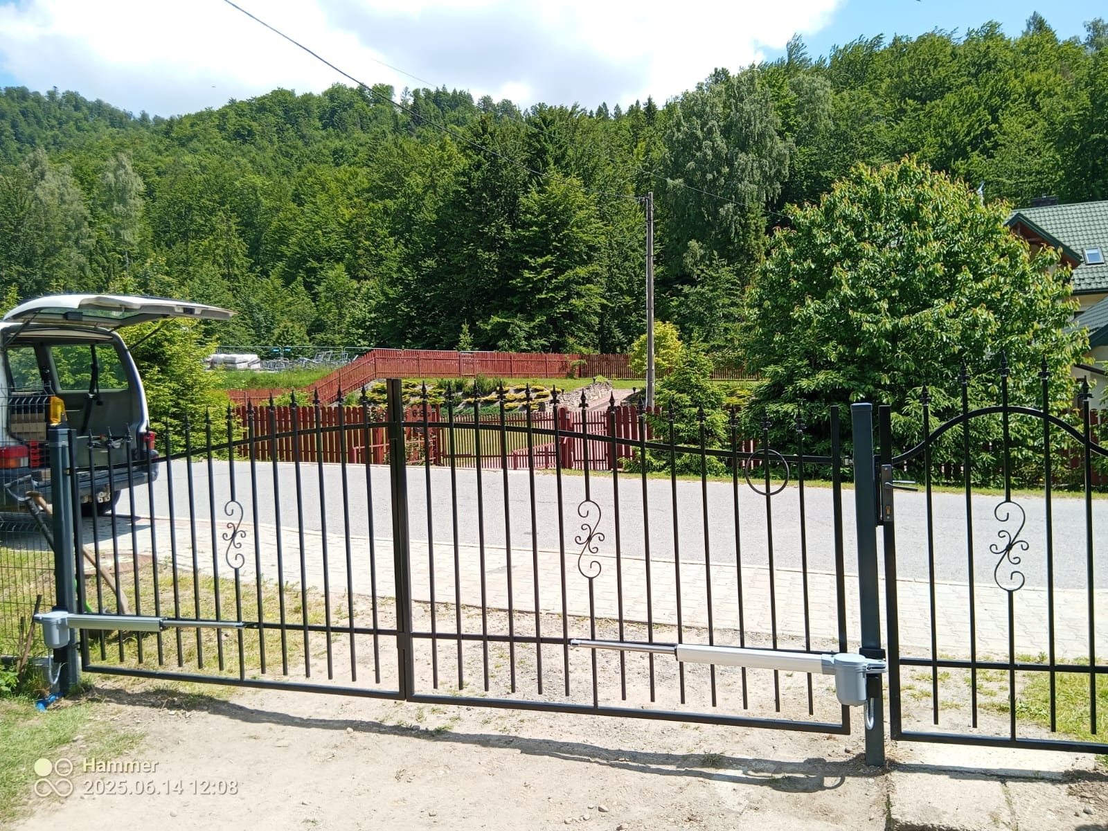 Czarna, metalowa brama wjazdowa z automatyką, widoczna na tle zielonego wzgórza i częściowo zaparkowanego samochodu dostawczego z otwartym bagażnikiem.