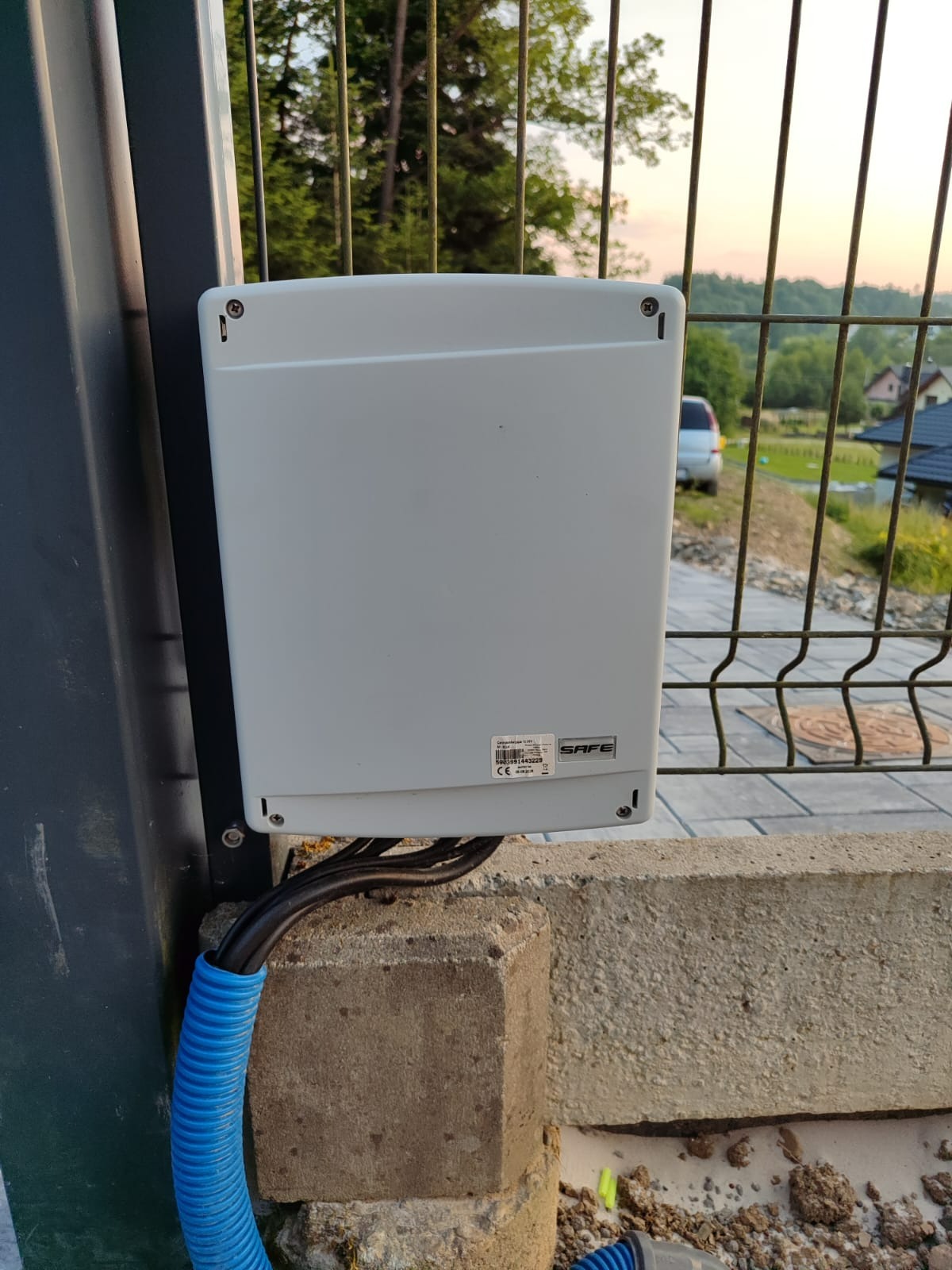 Szara obudowa sterownika bramy SAFE, zamocowana na słupku ogrodzenia, z widocznymi kablami w peszlu, na tle posesji i drzew w letni dzień.