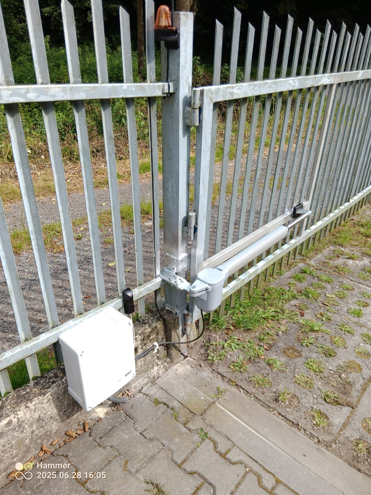 Szara, metalowa brama z zamontowaną automatyką, widoczny napęd liniowy, lampa ostrzegawcza i skrzynka sterująca, na tle zieleni.