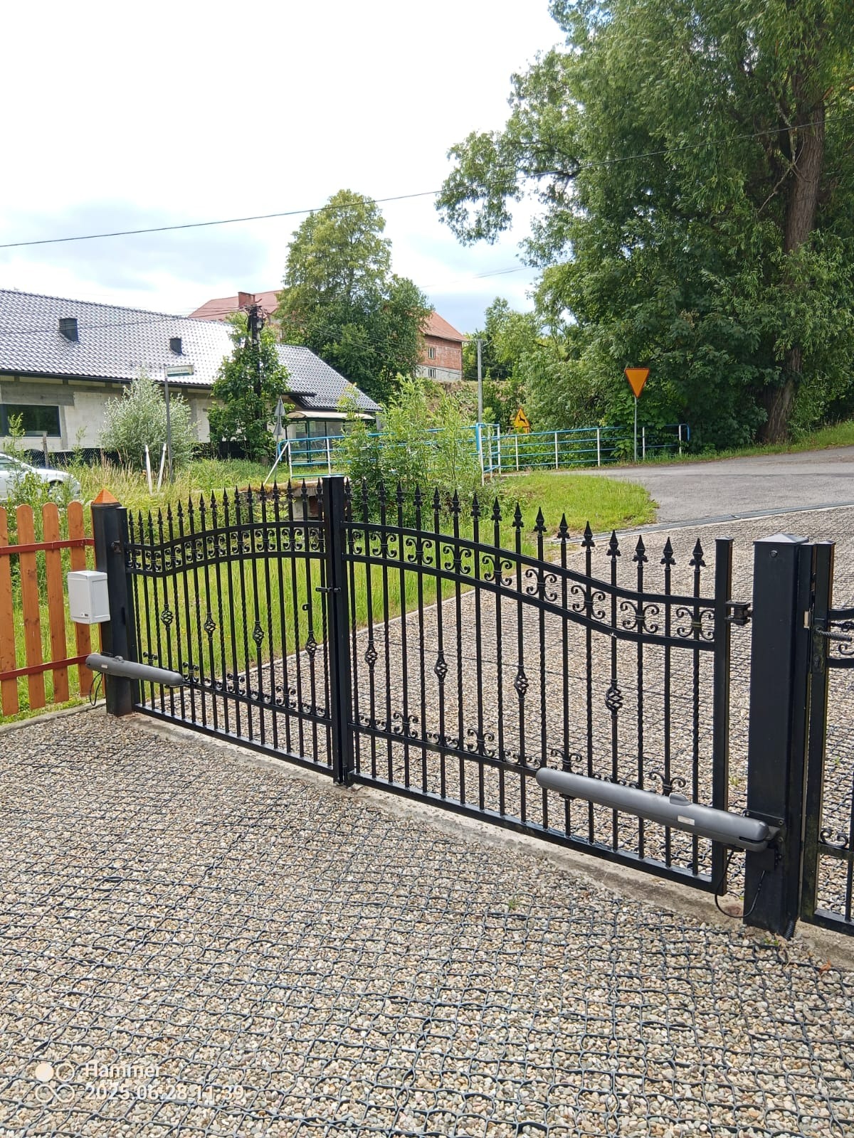Czarna, metalowa brama wjazdowa z automatyką na posesji z kamienistym podjazdem. W tle dom i zieleń. Widoczne elementy systemu automatycznego otwierania.