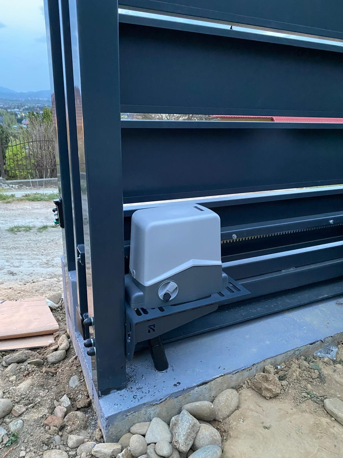 Szara automatyka do bramy przesuwnej, zamontowana na betonowej podstawie. Widoczne elementy konstrukcyjne bramy i tło z zielenią oraz górami.