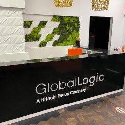 OKLEJWSZYSTKO.PL - Recepcja biura GlobalLogic z logo na czarnym blacie, zieloną ścianą z mchu i designerskimi lampami. Parkiet imitujący drewno w heksagonalnym wzorze.