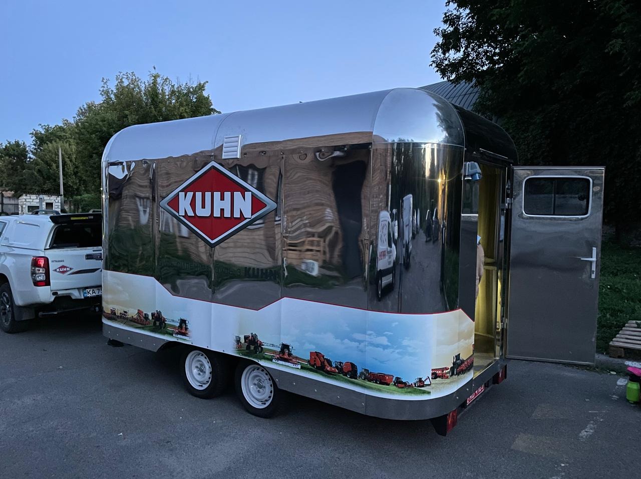 Oklejona przyczepa z logo KUHN i grafiką maszyn rolniczych na tle krajobrazu, zaparkowana obok białego pickupa. Drzwi przyczepy są otwarte.