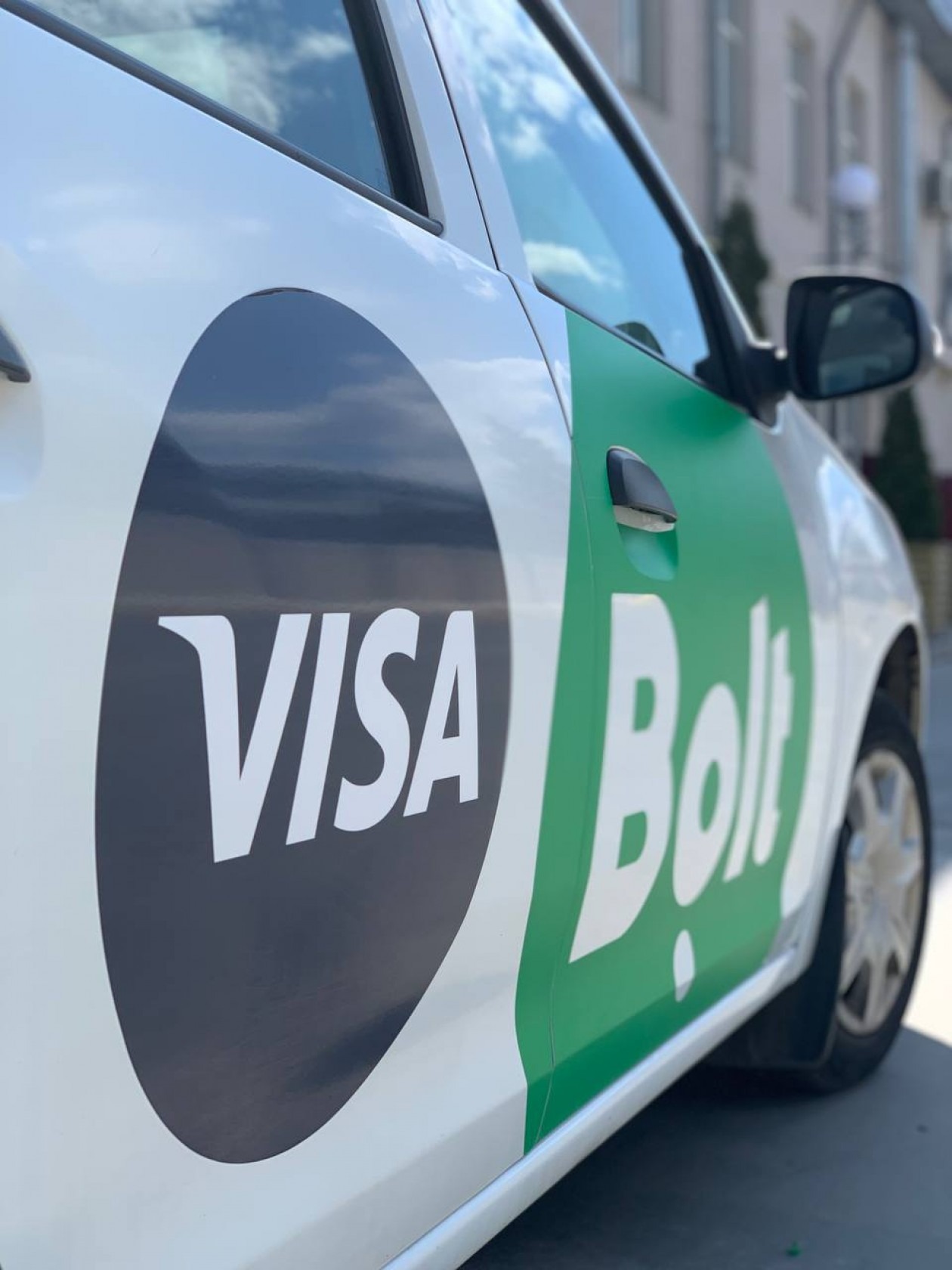 Oklejony bok białego auta z logotypami VISA i Bolt w czarno-zielonej kolorystyce. Widoczne odbicie chmur w lakierze. Perspektywa z dołu.
