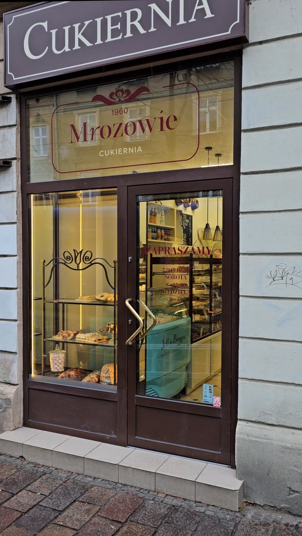 Witryna cukierni Mrozowie z szyldem i napisem 'Zapraszam'. Wnętrze z półkami pełnymi ciast i deserów, widoczna lodówka na słodkości.