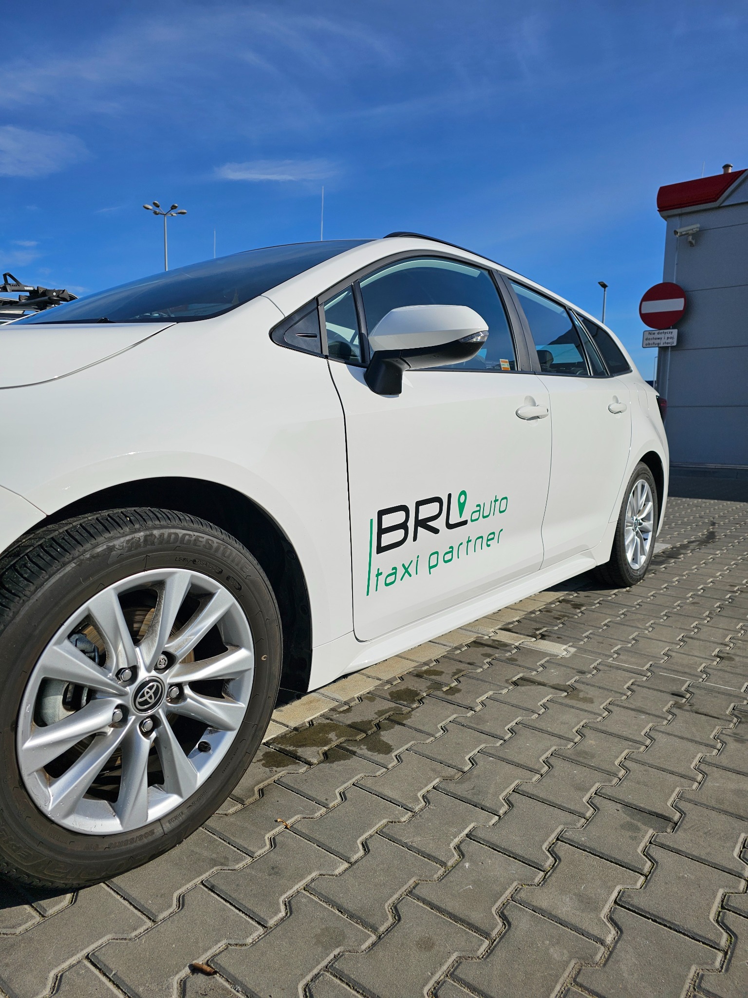 Biała Toyota z zielonym logo 'BRL auto taxi partner' na boku, zaparkowana na kostce brukowej przed budynkiem w słoneczny dzień. Oznakowanie pojazdu.