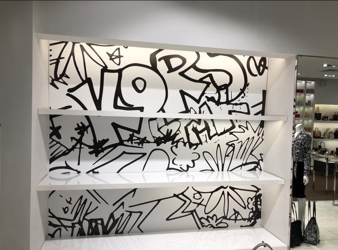 Biała półka sklepowa z czarnymi, abstrakcyjnymi grafikami na ścianach, tworzącymi unikalny wzór. Oświetlone, błyszczące półki.