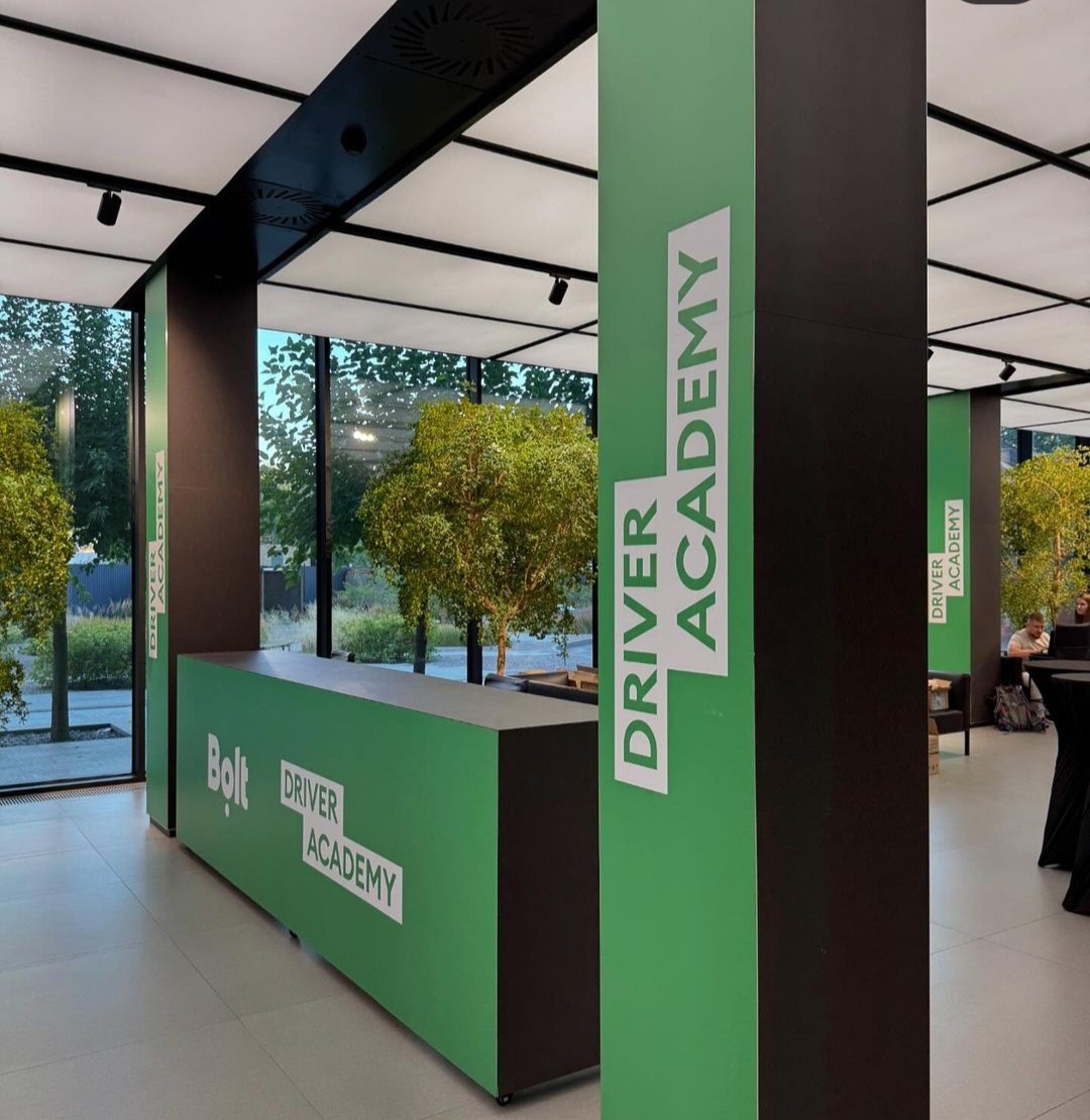 Wnętrze biura Bolt Driver Academy z zielonymi elementami identyfikacji wizualnej, widoczne logo i nazwa akademii na ladzie recepcyjnej i filarach.
