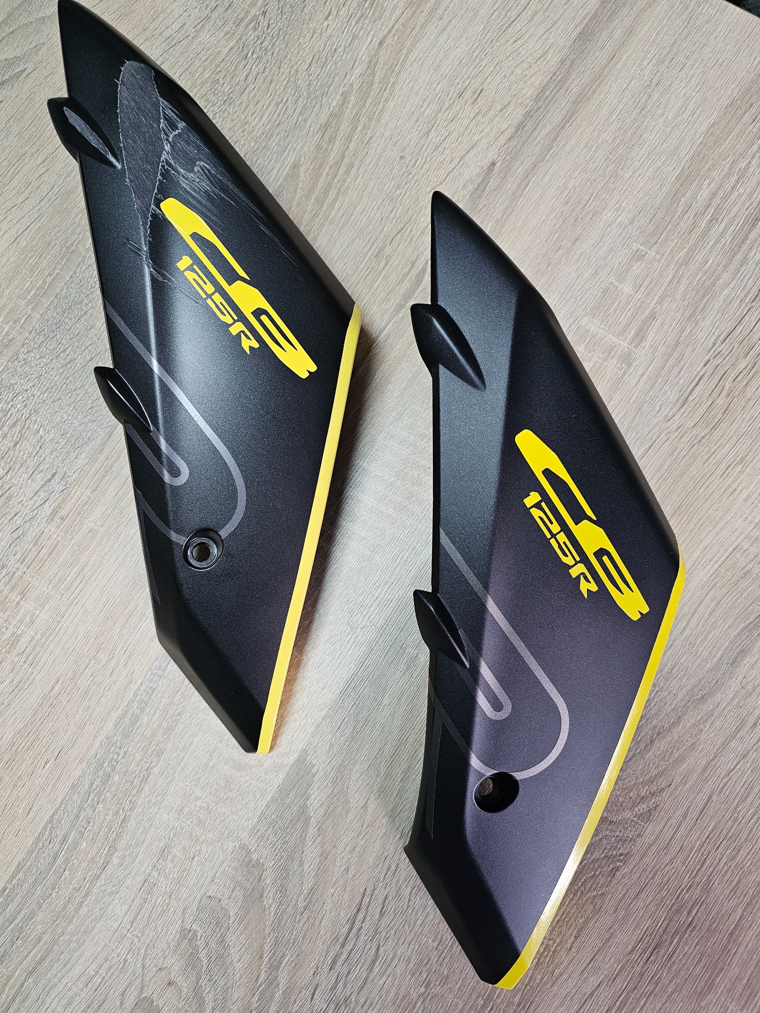 Dwa czarne elementy motocyklowe z żółtymi akcentami i napisem 'CB 125R', jeden z widocznymi zarysowaniami, leżą na drewnianej powierzchni.