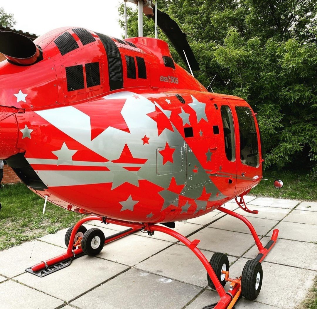 Czerwony helikopter Bell 505 z srebrnymi gwiazdami na karoserii, stojący na betonowej nawierzchni, z zielonymi drzewami w tle. Detal oklejania.