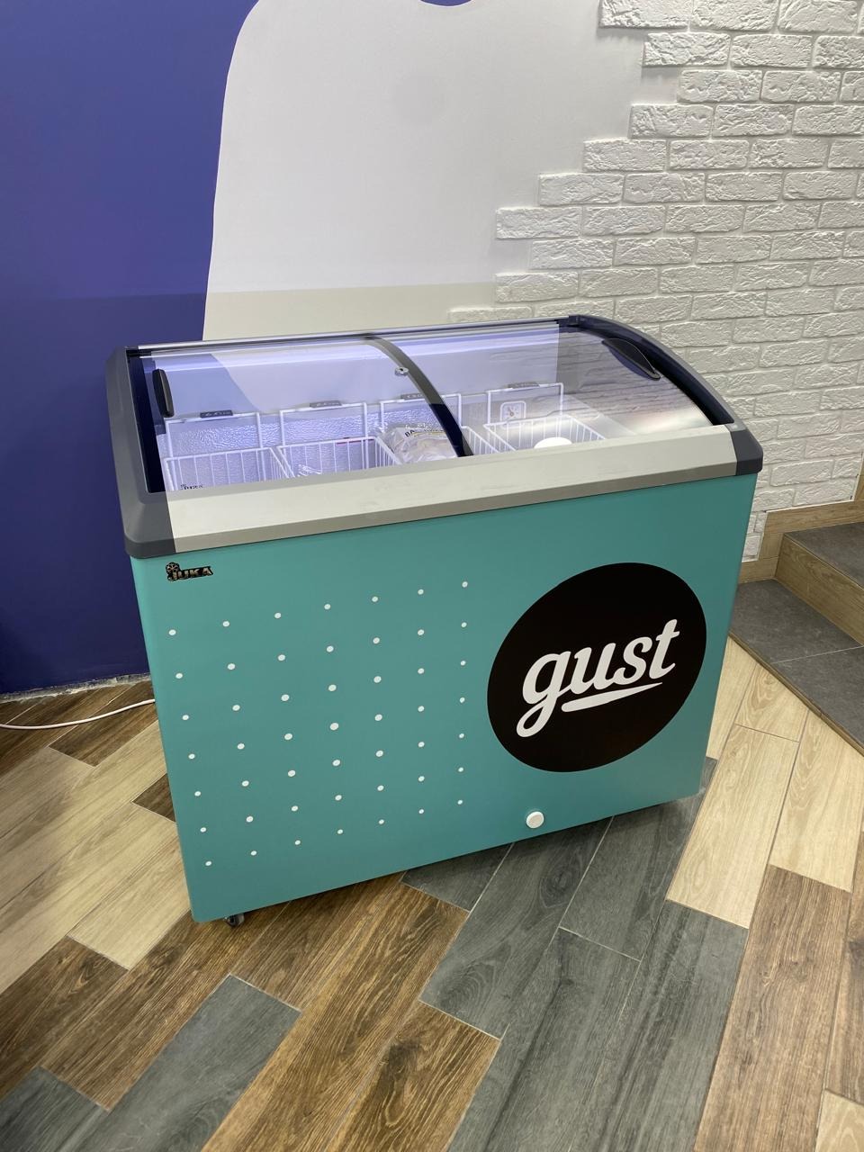 Turkusowa lodówka z logo 'gust' na tle białej cegły i niebieskiej ściany. Widoczna podłoga z paneli. Chłodnia z lodami w sklepie.