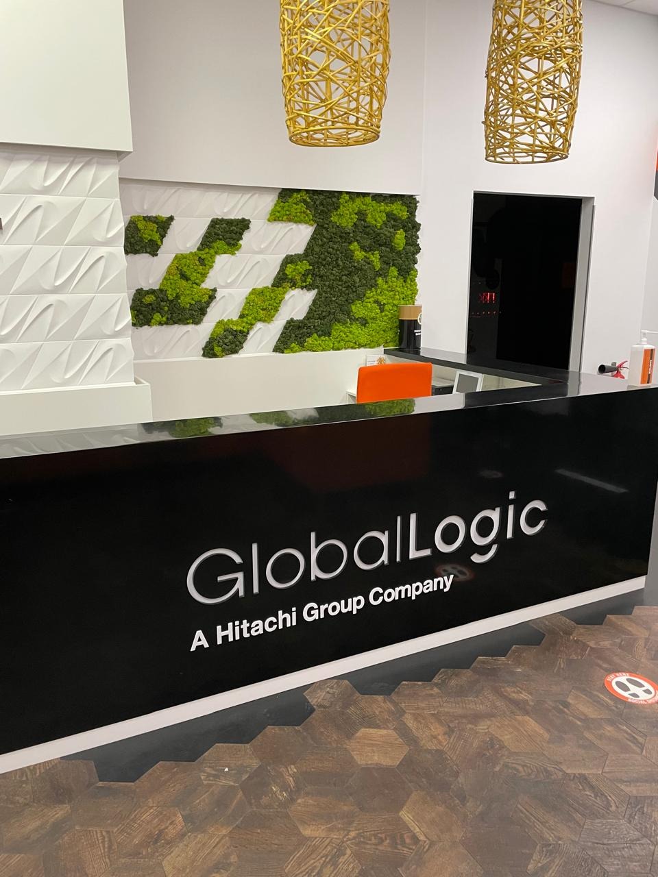 Recepcja biura GlobalLogic z logo na czarnym blacie, zieloną ścianą z mchu i designerskimi lampami. Parkiet imitujący drewno w heksagonalnym wzorze.