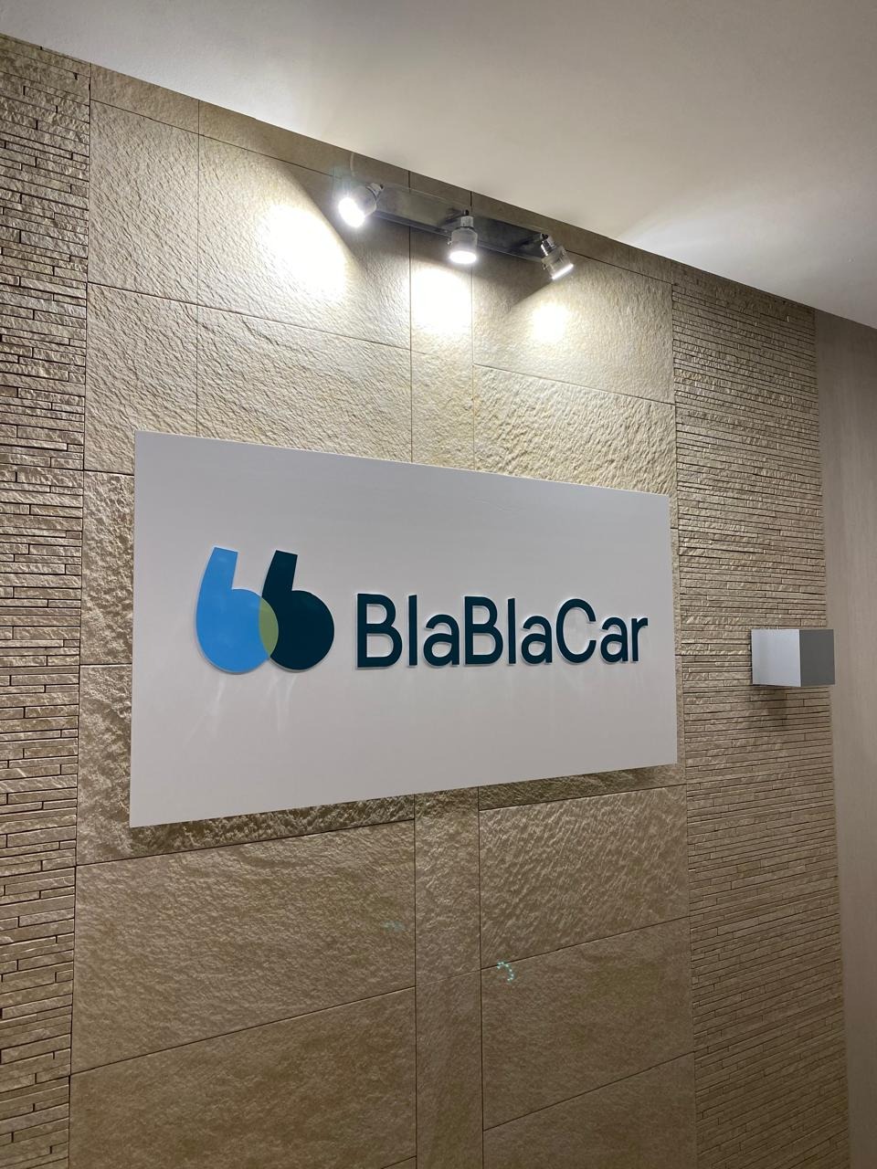 Biały szyld z logo BlaBlaCar na teksturowanej ścianie z oświetleniem punktowym. Logo w odcieniach niebieskiego i zieleni. Minimalistyczny design, nowoczesny wygląd.