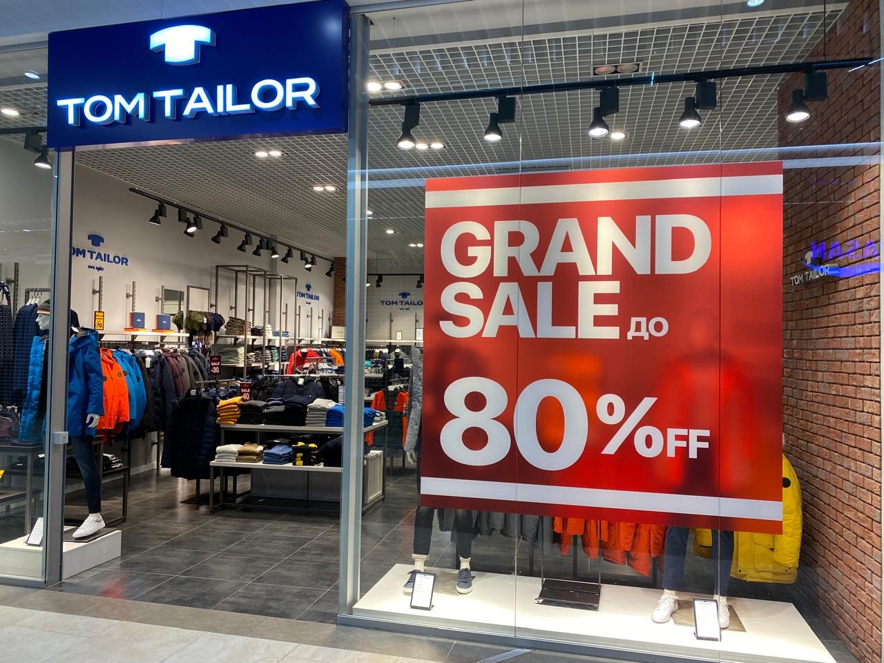 Wnętrze sklepu Tom Tailor z wywieszką 'GRAND SALE do 80% OFF' na witrynie. Widoczne ubrania na wieszakach i manekin w niebieskiej kurtce.