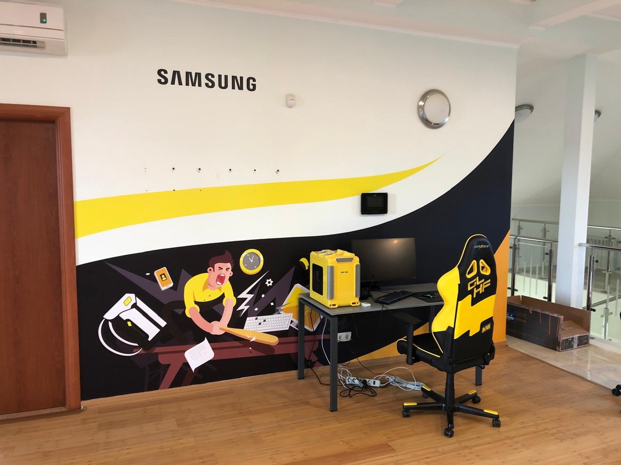 Ściana z naklejką Samsung i motywem gracza, biurko z komputerem i żółto-czarne krzesło gamingowe w przestronnym wnętrzu. Dekoracja biura w stylu gamingowym.