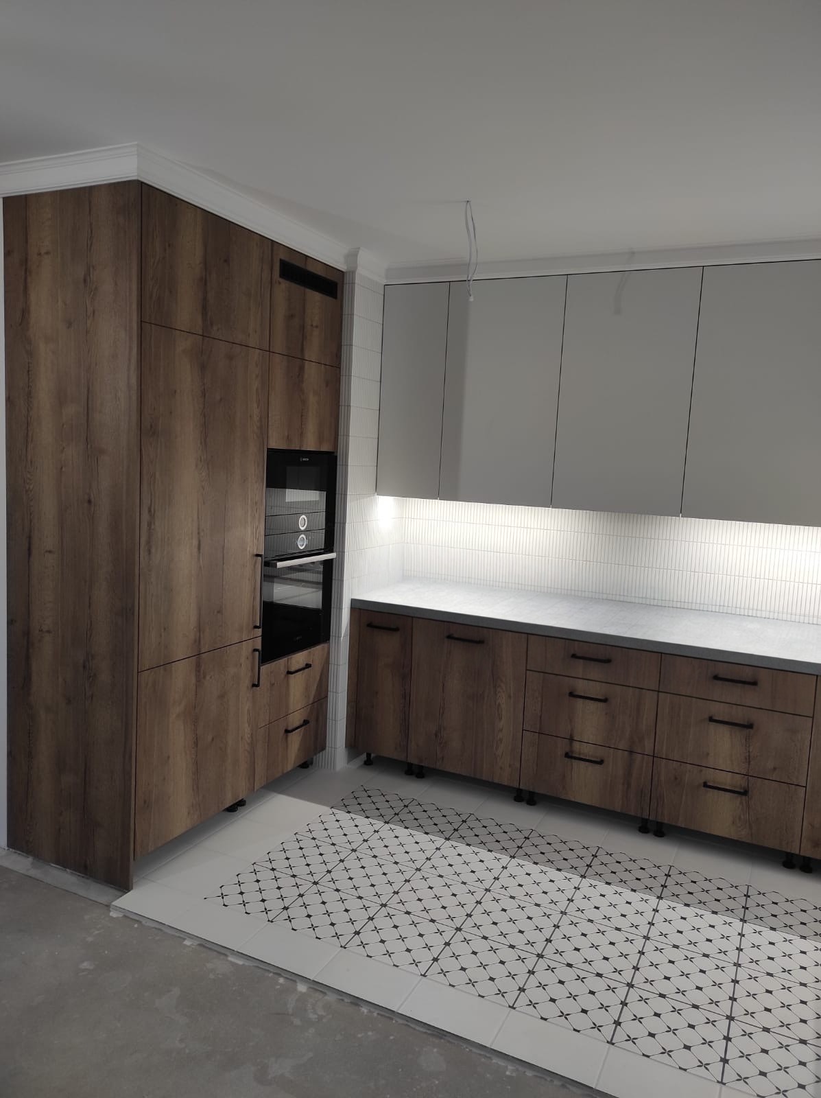 Nowoczesna kuchnia z drewnopodobnymi szafkami dolnymi i szarymi górnymi, oświetleniem LED pod szafkami i wzorzystą podłogą, z piekarnikiem w zabudowie.