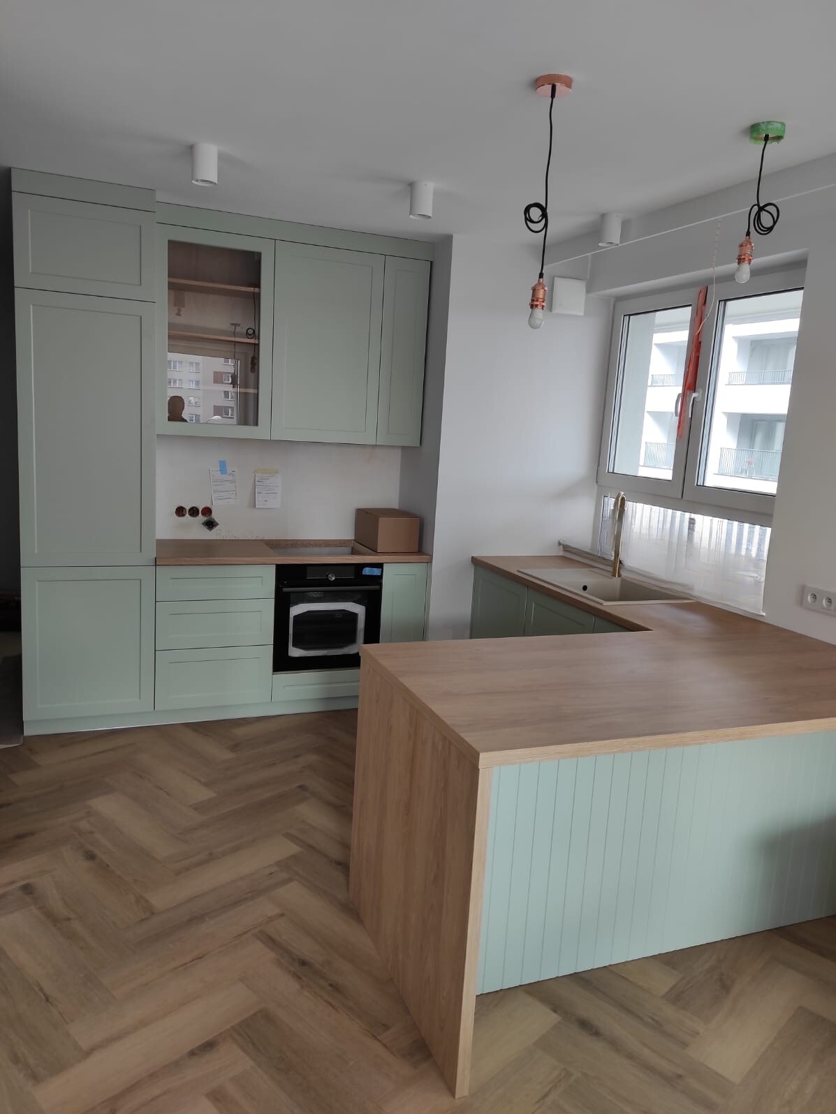 Jasnozielona kuchnia na wymiar z drewnianym blatem i podłogą w jodełkę. Nad blatem wiszą lampy z miedzianymi akcentami. Widok na okno z białymi ramami.