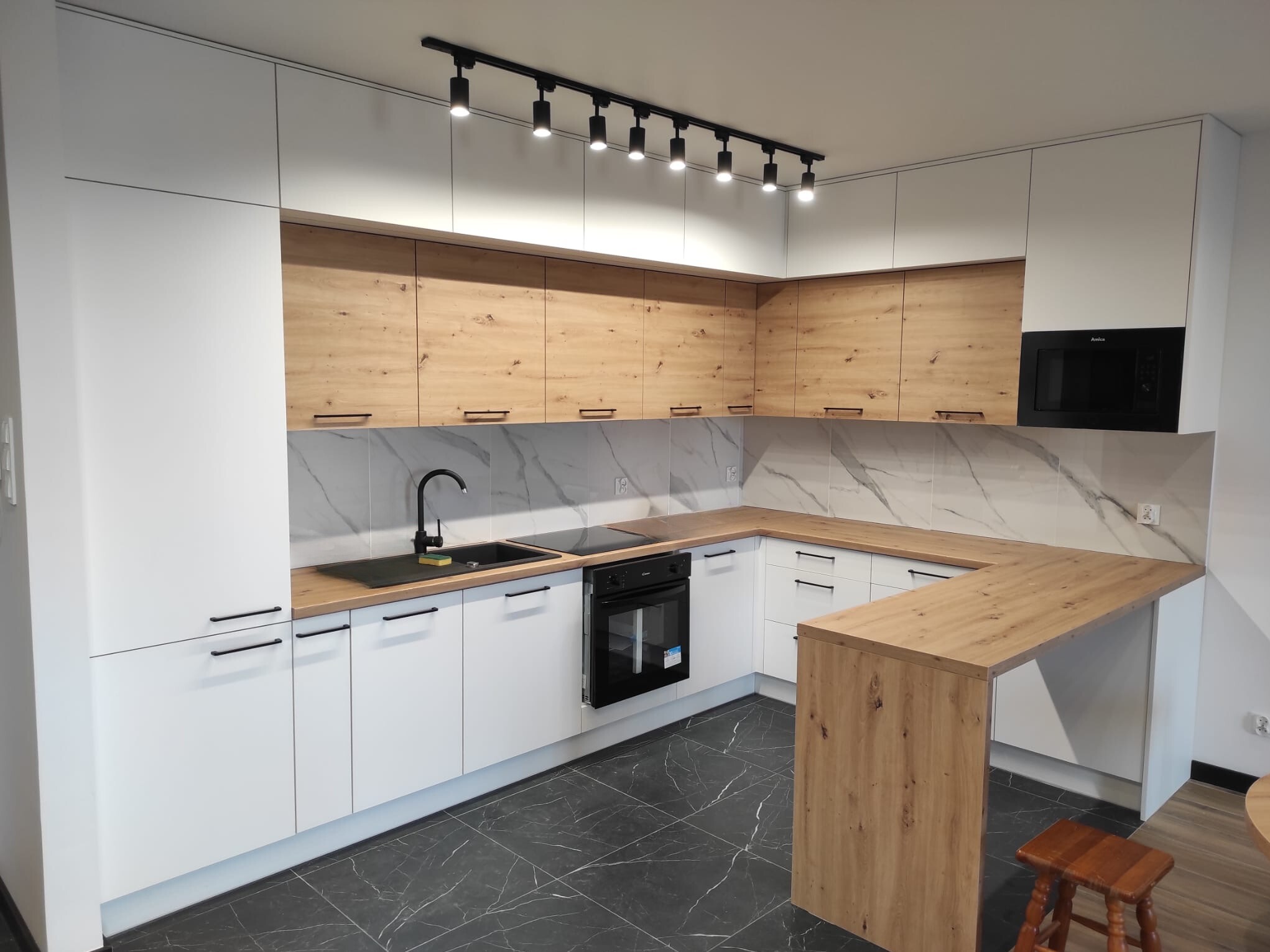 Nowoczesna kuchnia na wymiar z białymi frontami i drewnianymi akcentami, czarne uchwyty i oświetlenie szynowe, marmurowy backsplash, czarny zlew i płyta grzewcza.