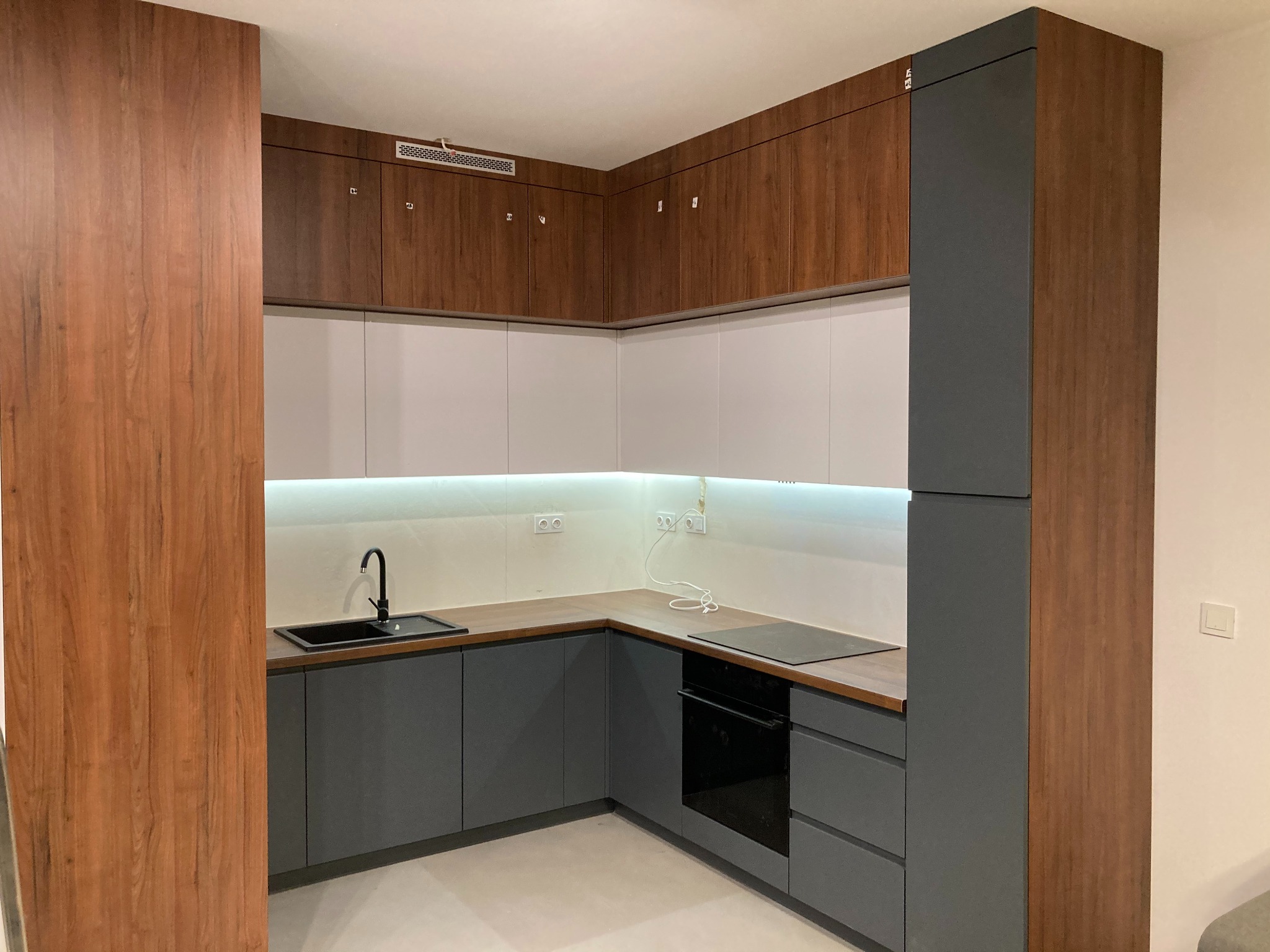 Nowoczesna kuchnia z szarymi szafkami dolnymi i drewnopodobnymi górnymi, z czarnym zlewem i płytą indukcyjną. Oświetlenie LED pod szafkami.