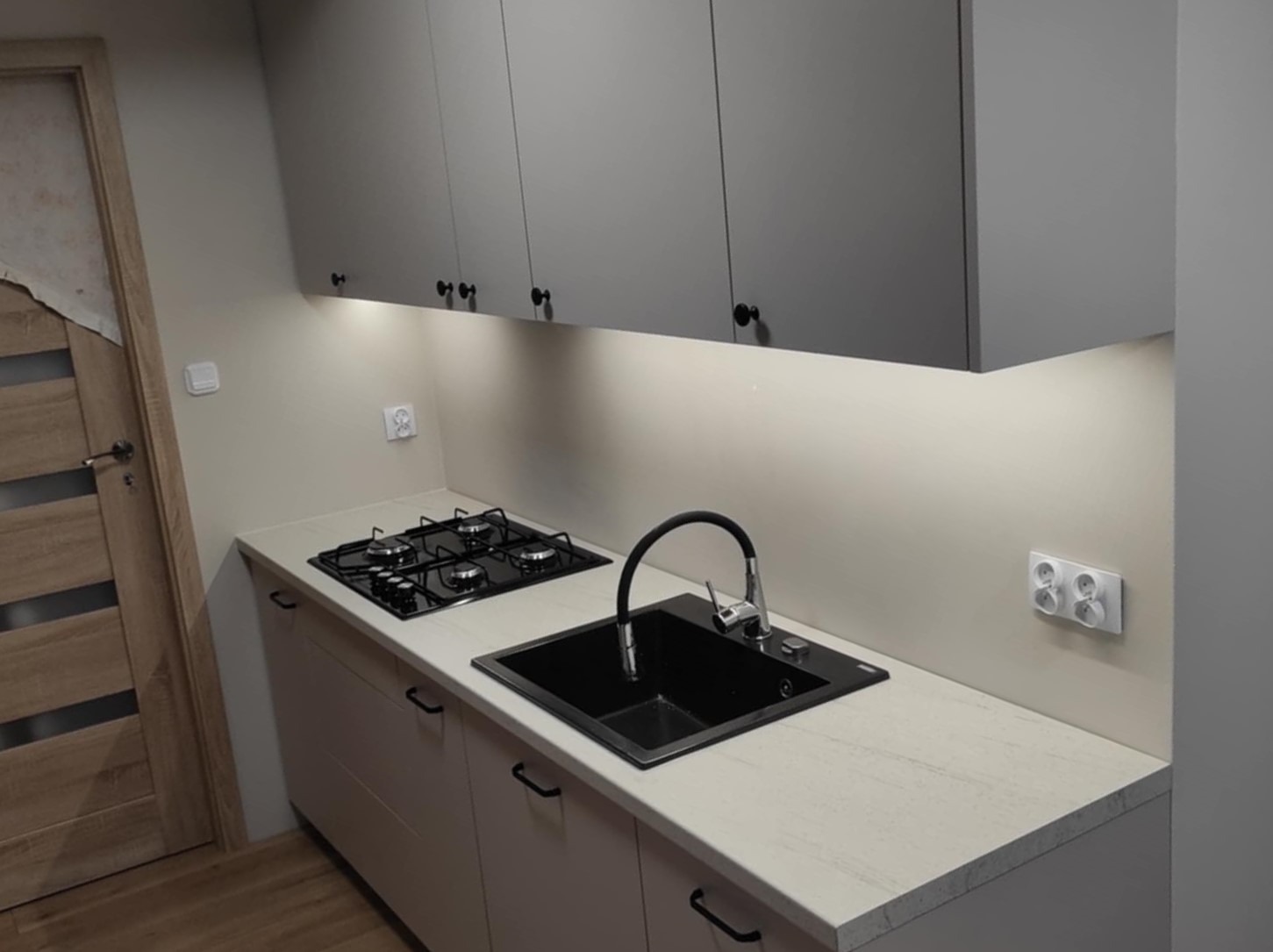 Szare szafki wiszące nad beżowym blatem z czarnym zlewem i kuchenką gazową. Jasne oświetlenie podszafkowe.  Ujęcie z niskiego kąta, widoczny fragment drzwi.