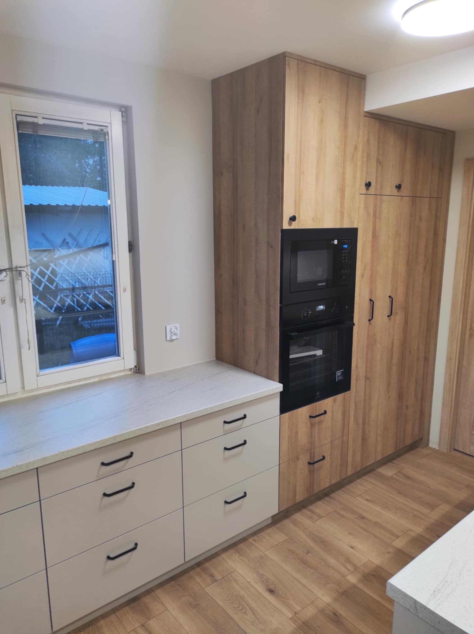 Jasna kuchnia na wymiar z drewnianymi szafkami, czarnym piekarnikiem i mikrofalówką w zabudowie. Blat w kolorze jasnego kamienia. Widok na okno.